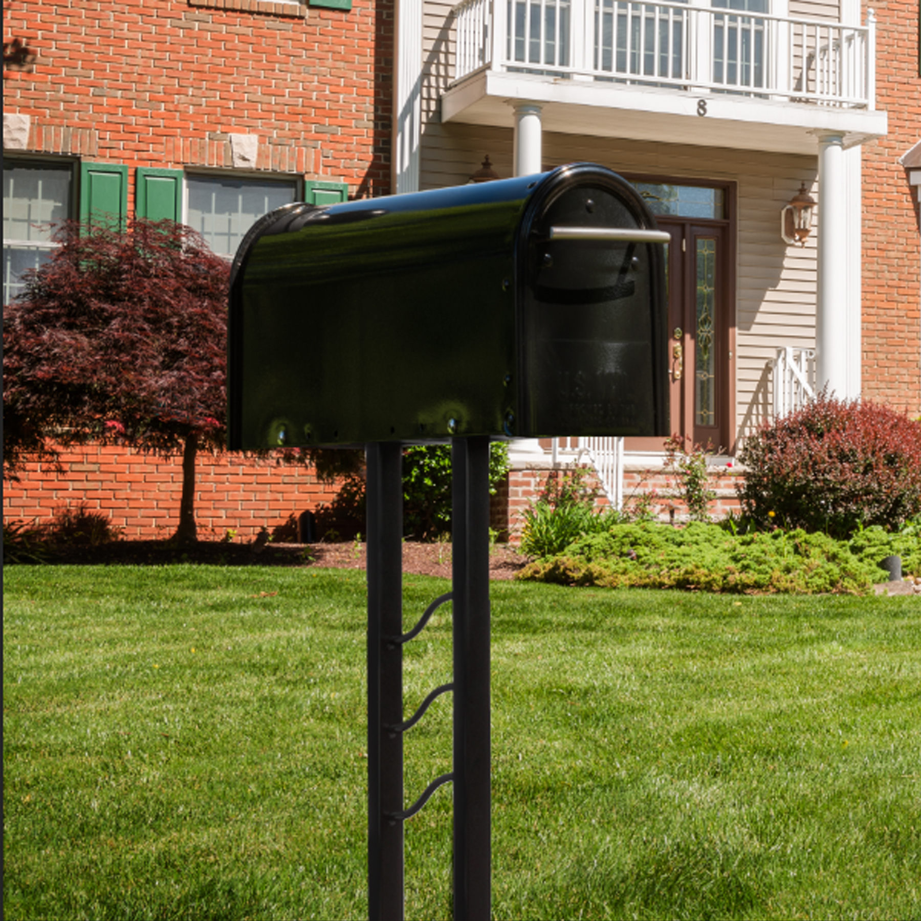 Green curved-top mailbox on a black metal post in a front yard, with a brick house and white porch in the background.