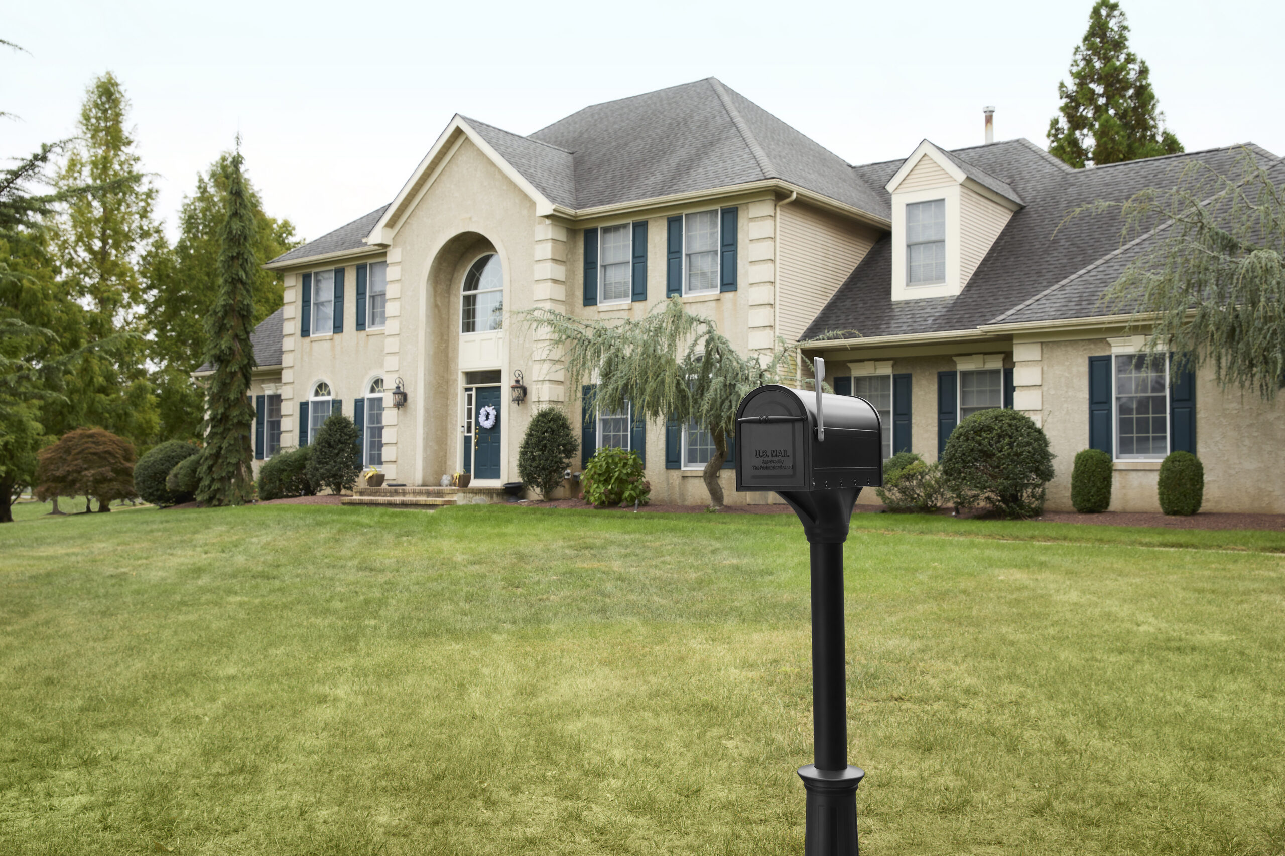 Front yard view of a large beige suburban house with blue shutters and a black mailbox in the foreground.