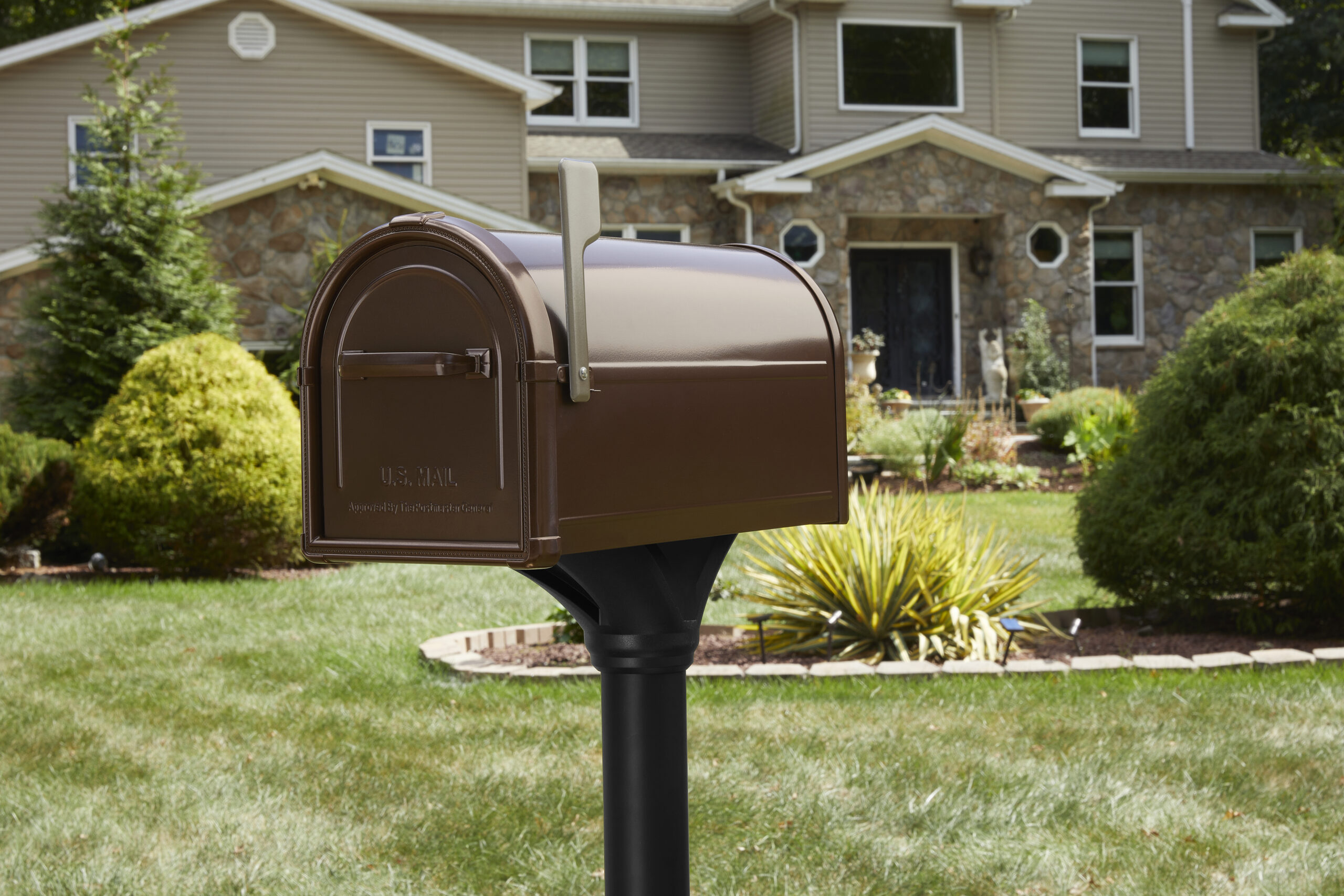 Brown curbside mailbox with a rounded top on a black post in a manicured front yard of a suburban house with stone accents behind it.