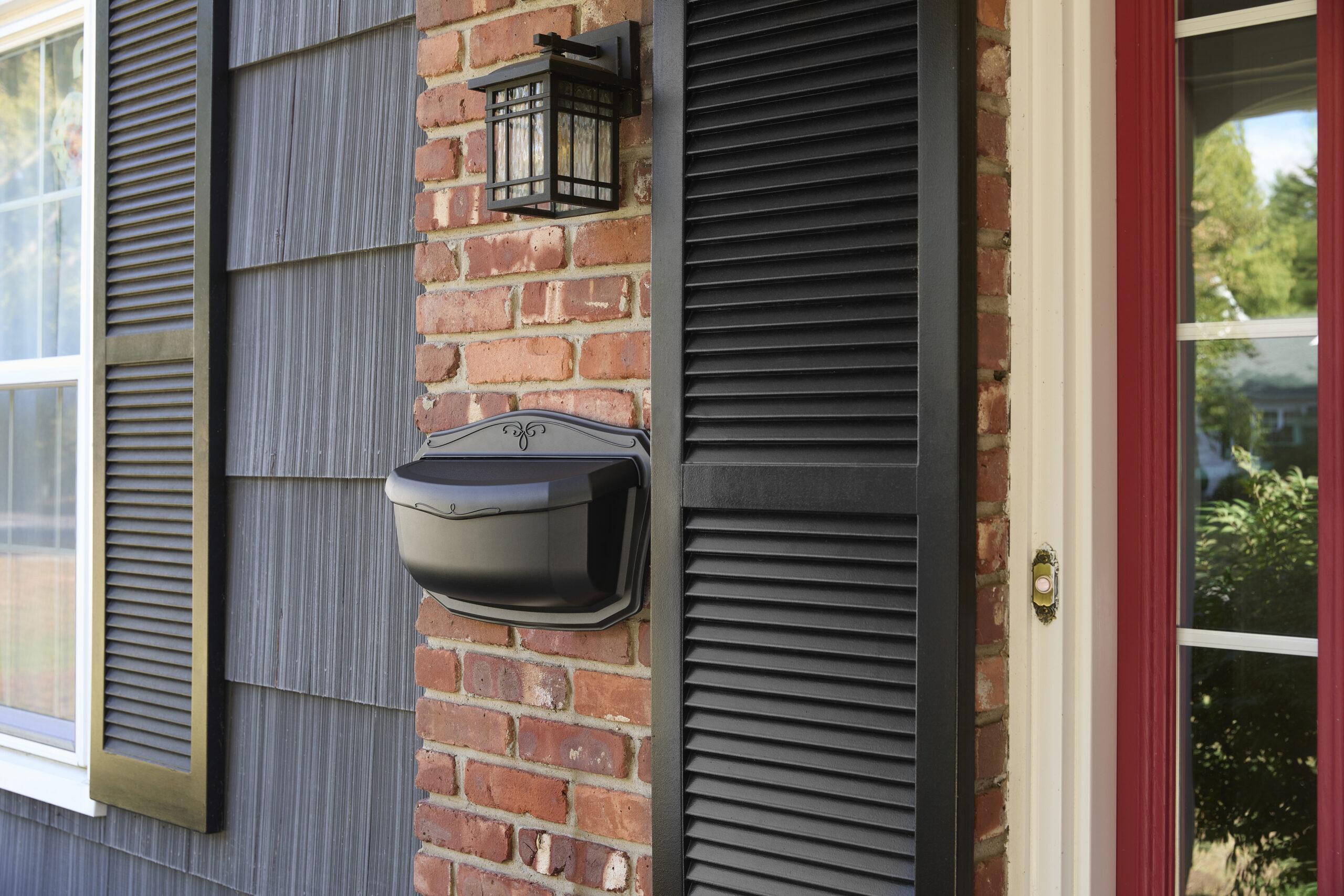 Front porch area with a black mailbox on a brick wall, black shutters, a lantern above, and a red-framed door on the right.