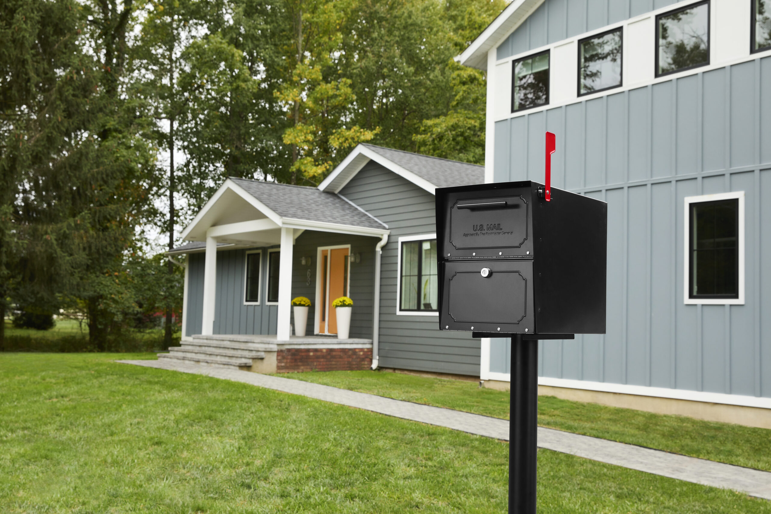 Black curbside mailbox with red flag mounted on a post in front of a modern blue house with a porch and yellow potted plants.