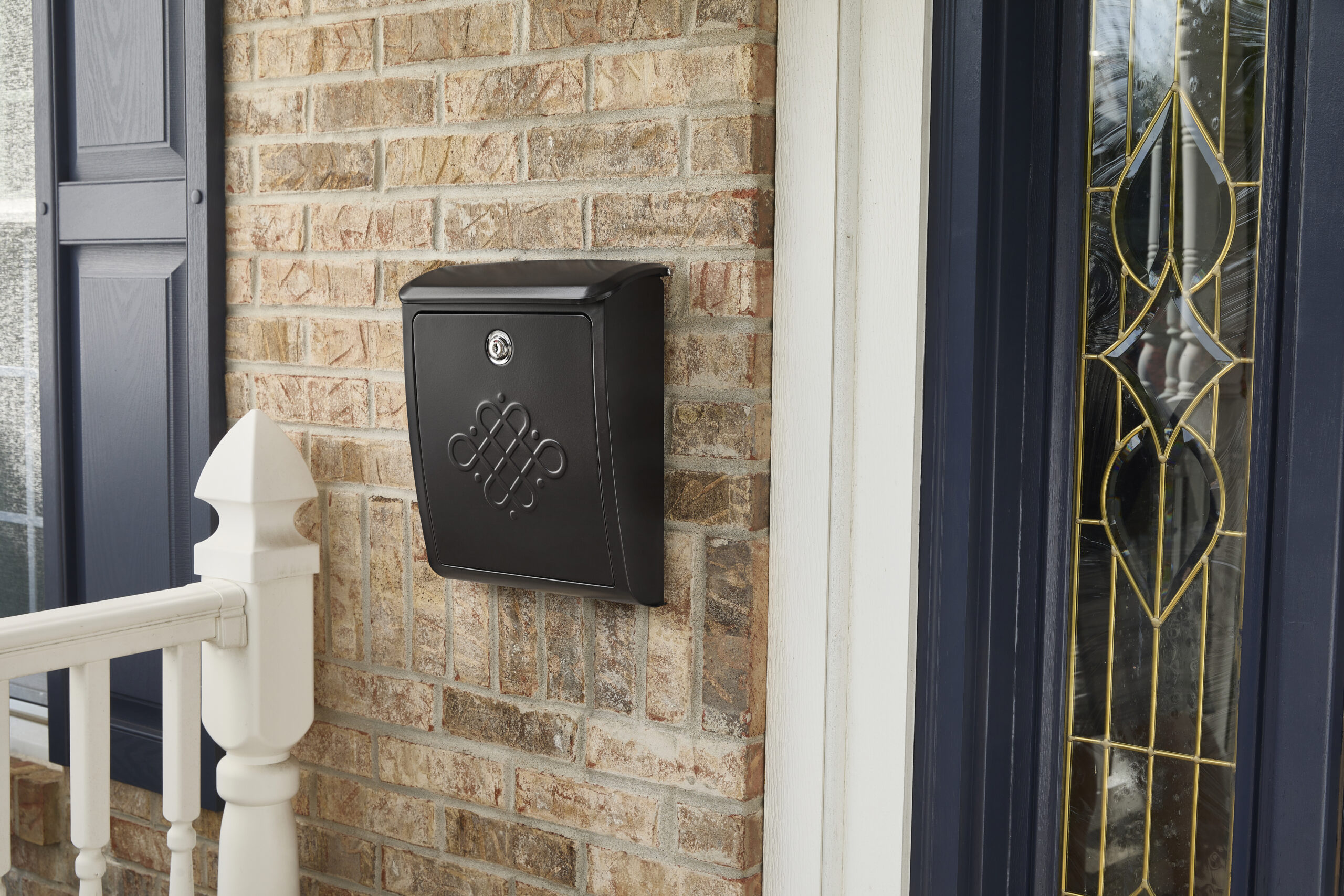 Black wall mailbox with decorative embossing mounted on a brick exterior beside a front door with stained-glass panels.