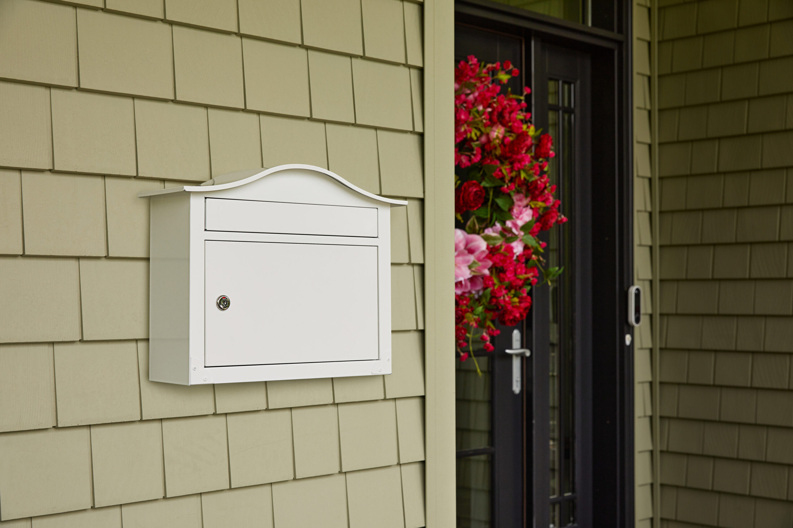 White wall-mounted mailbox with a curved top beside a front door wrapped in red and pink floral arrangements on a beige siding wall.