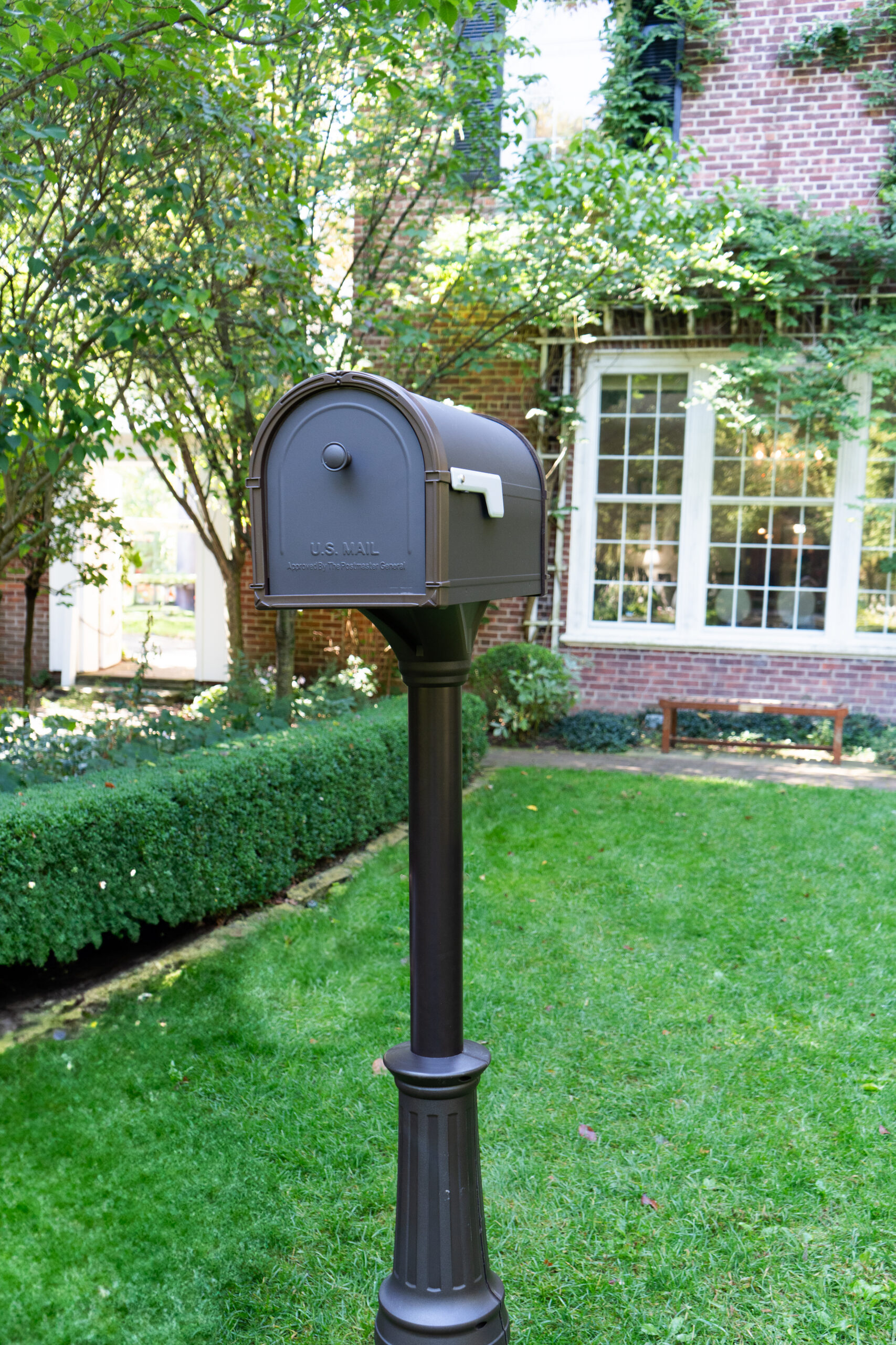 Metal U.S. Mail mailbox on a tall black post in a neatly mowed front yard with a brick house in the background.
