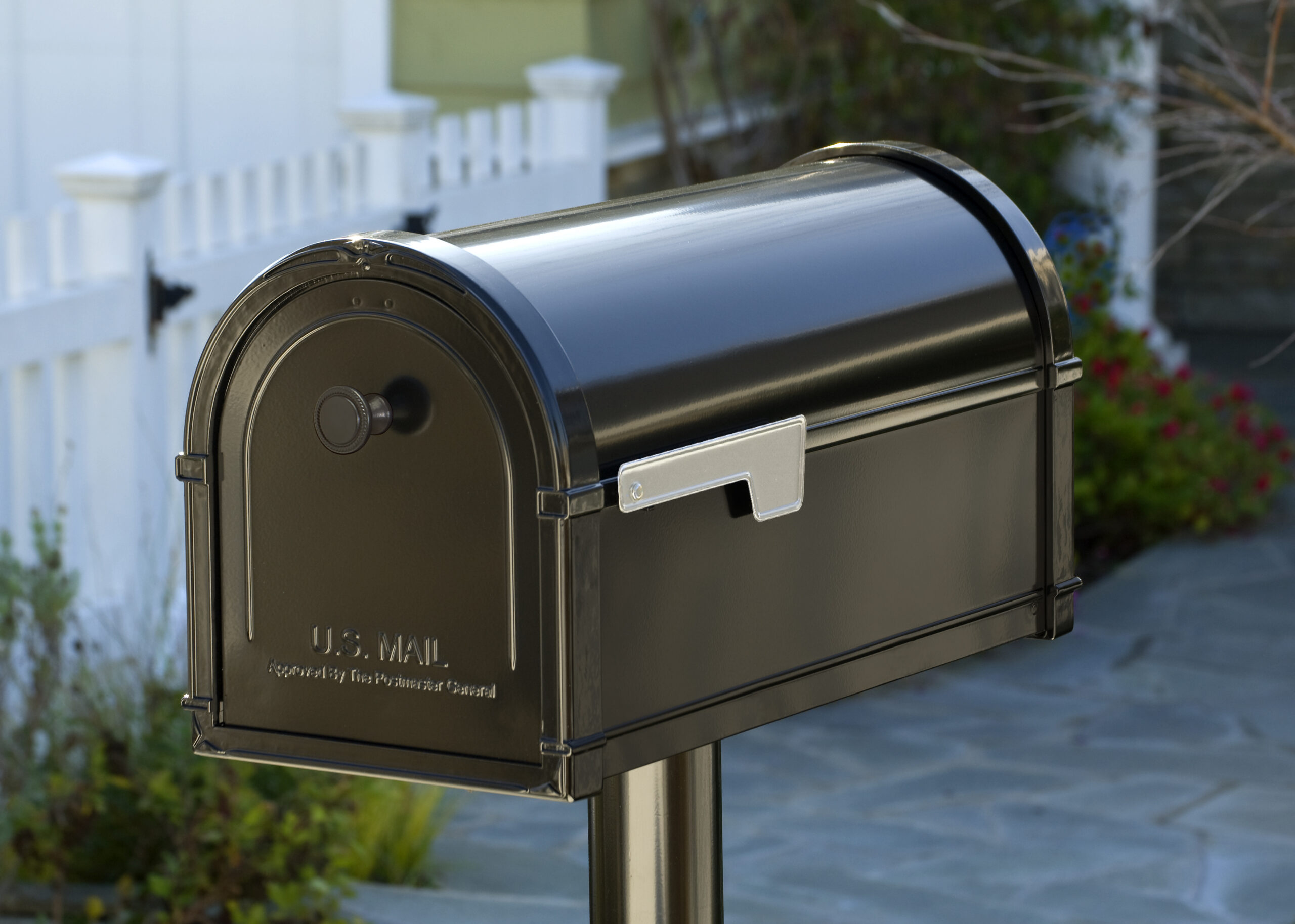 Black roll-top U.S. Mail mailbox on a post by a suburban walkway; stamped 'U.S. MAIL' on the door.