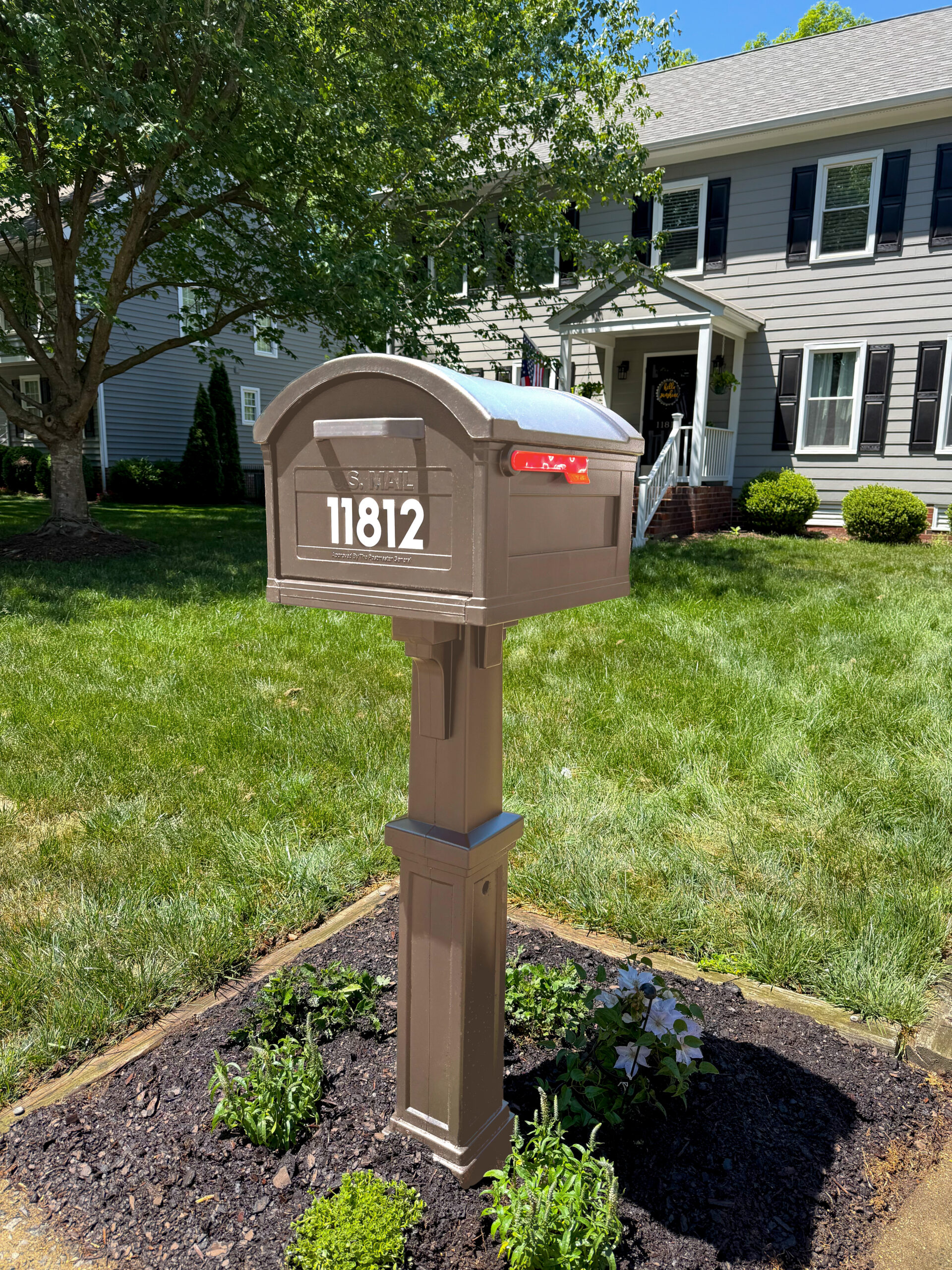 Brown curbside mailbox with curved top in a landscaped front yard, in front of a gray two-story house with black shutters; house number 11812 visible.