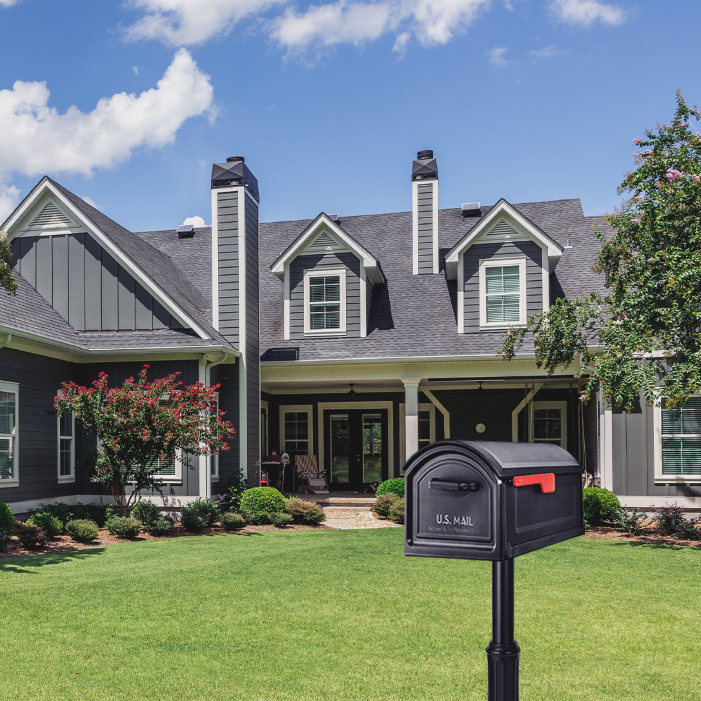 Front yard of a large gray two-story house with dormer windows and a black mailbox in the foreground on a well-kept lawn.