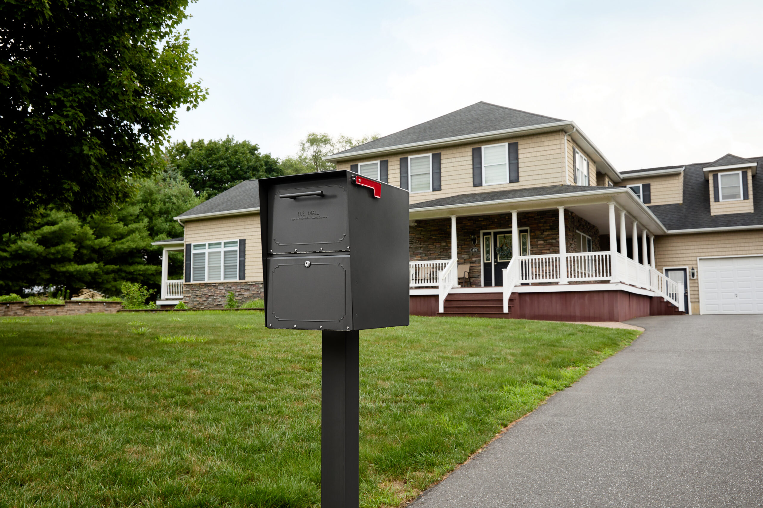Gray curbside mailbox on a post in a front yard with a two-story beige house and white porch in the background.