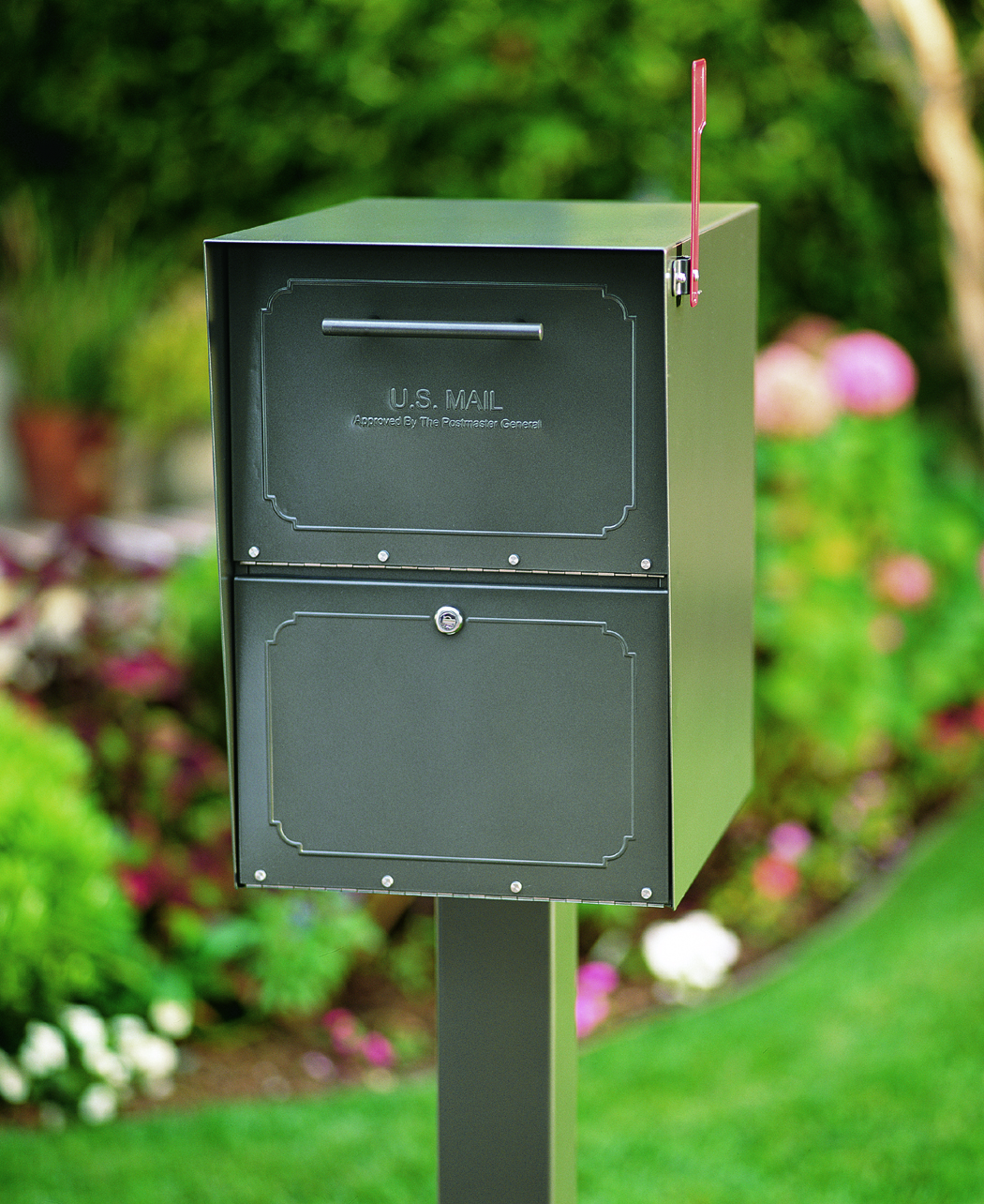 Green double-compartment U.S. Mail mailbox on a post in a landscaped garden; red flag raised on the right side.