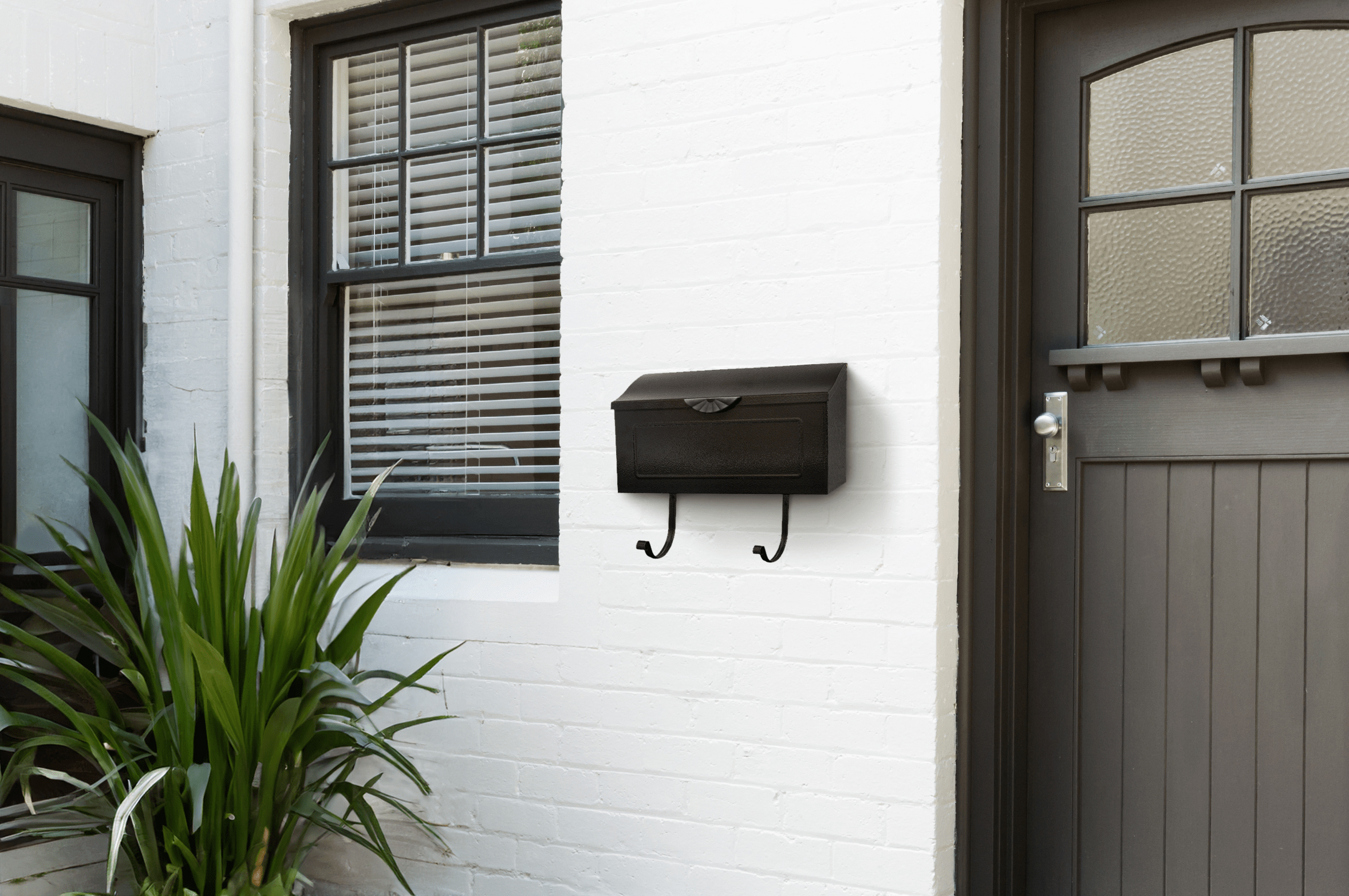 Black wall mailbox with two curved hooks mounted on a white brick wall beside a window and door.