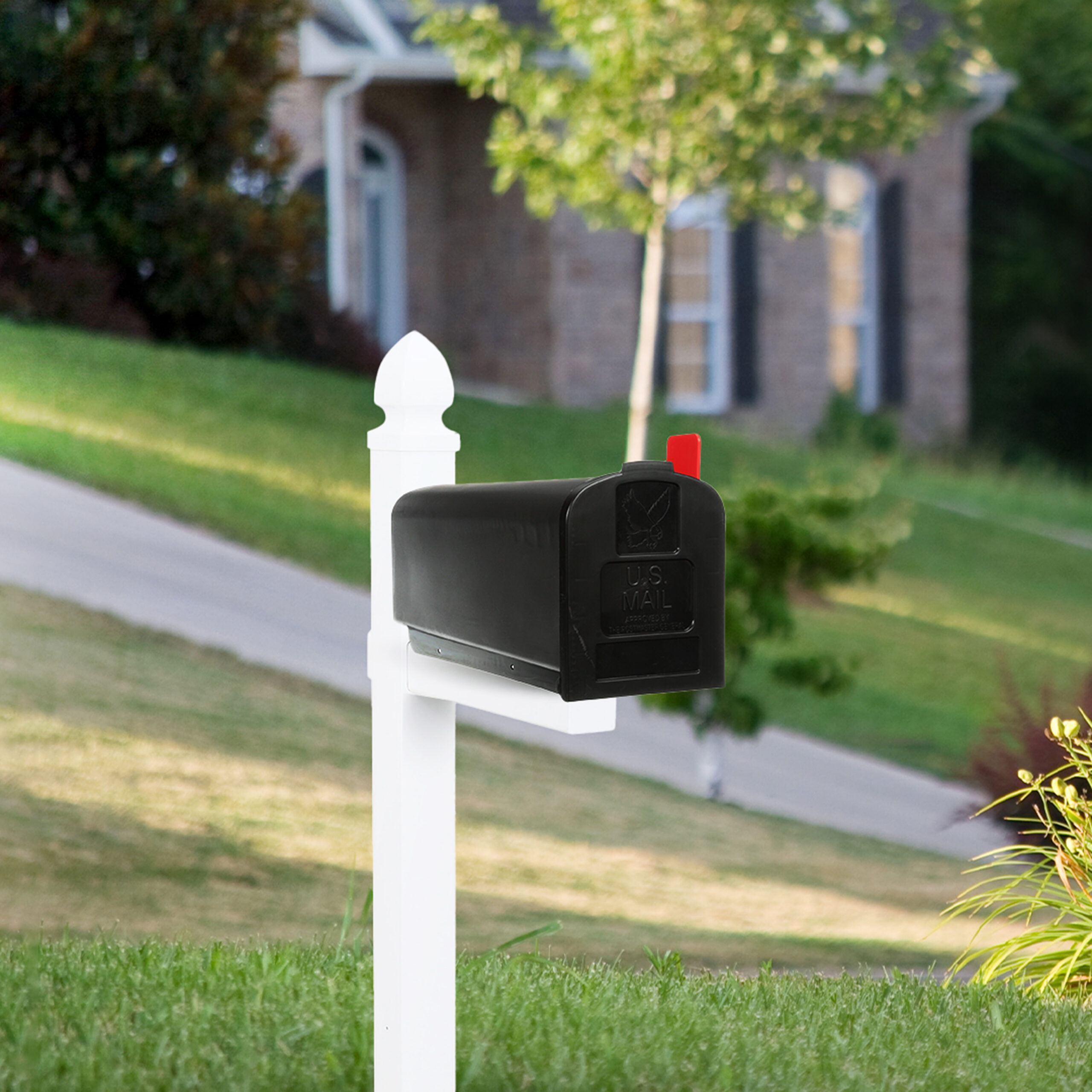 Black curbside mailbox on a white post with red flag raised in a suburban yard, with a house and green lawn in the background.