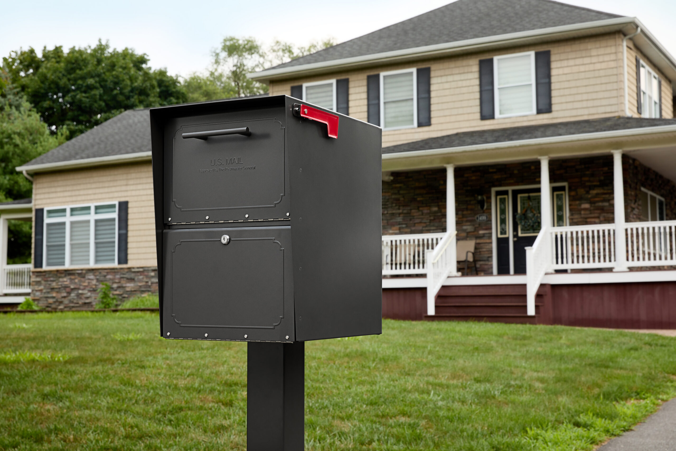 Gray two-compartment mailbox on a black post in a suburban front yard, red flag raised, in front of a two-story house with a porch.