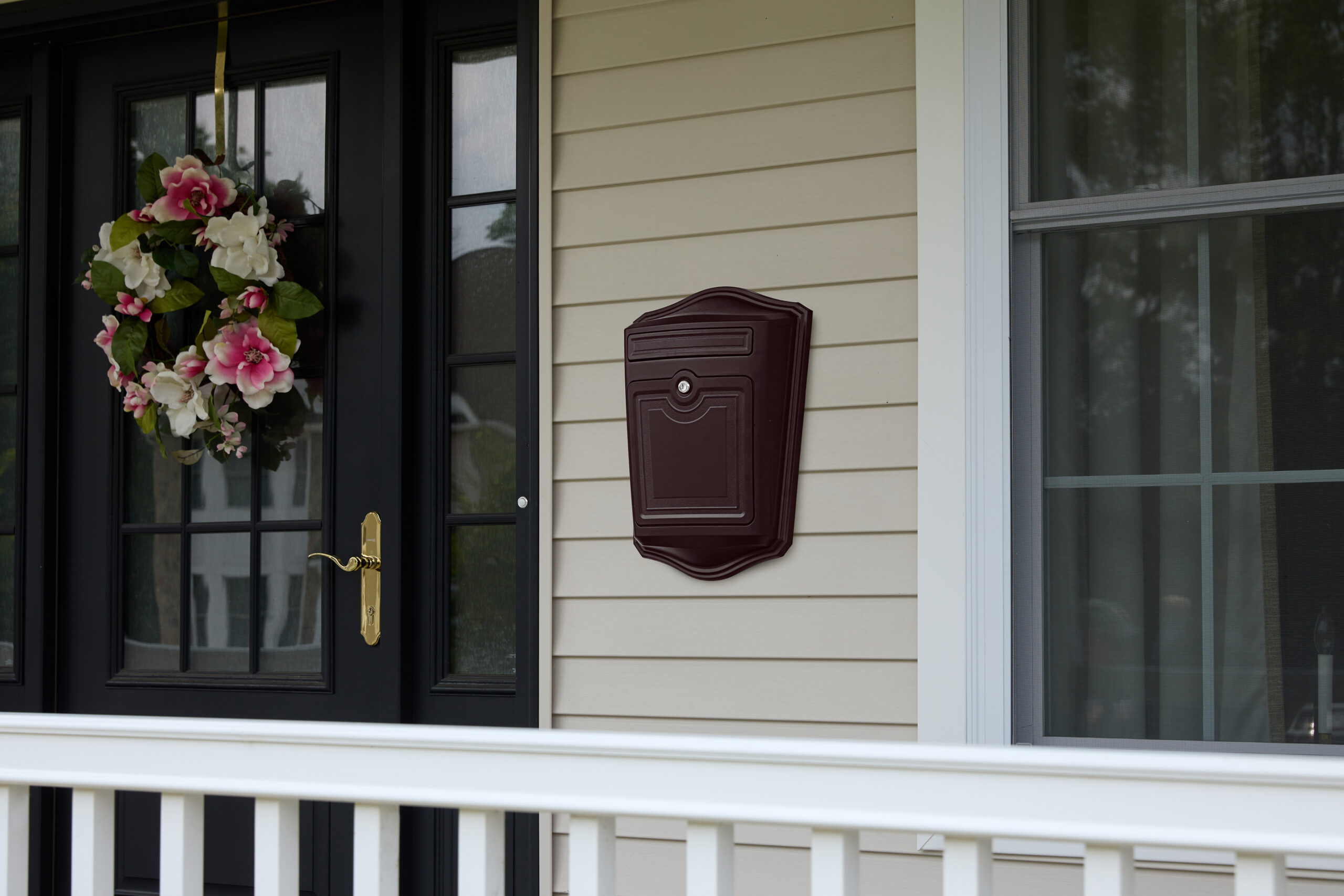 Front porch view featuring a black door with a pink and white floral wreath, beige siding, a maroon wall mailbox, and a white railing below.