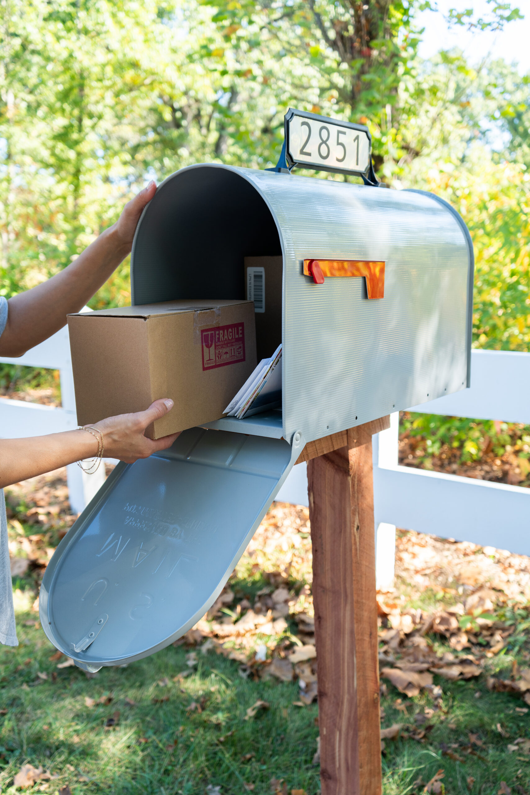 A person places a cardboard package into a curved metal curbside mailbox with the flag up and the number 2851 visible outdoors by a white fence.