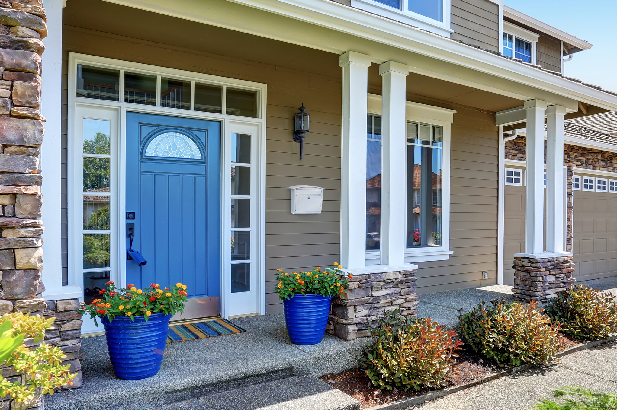 Blue front door with a semi-circular window sits on a beige house with white trim and stone accents, two blue planters with flowers flank the entryway.