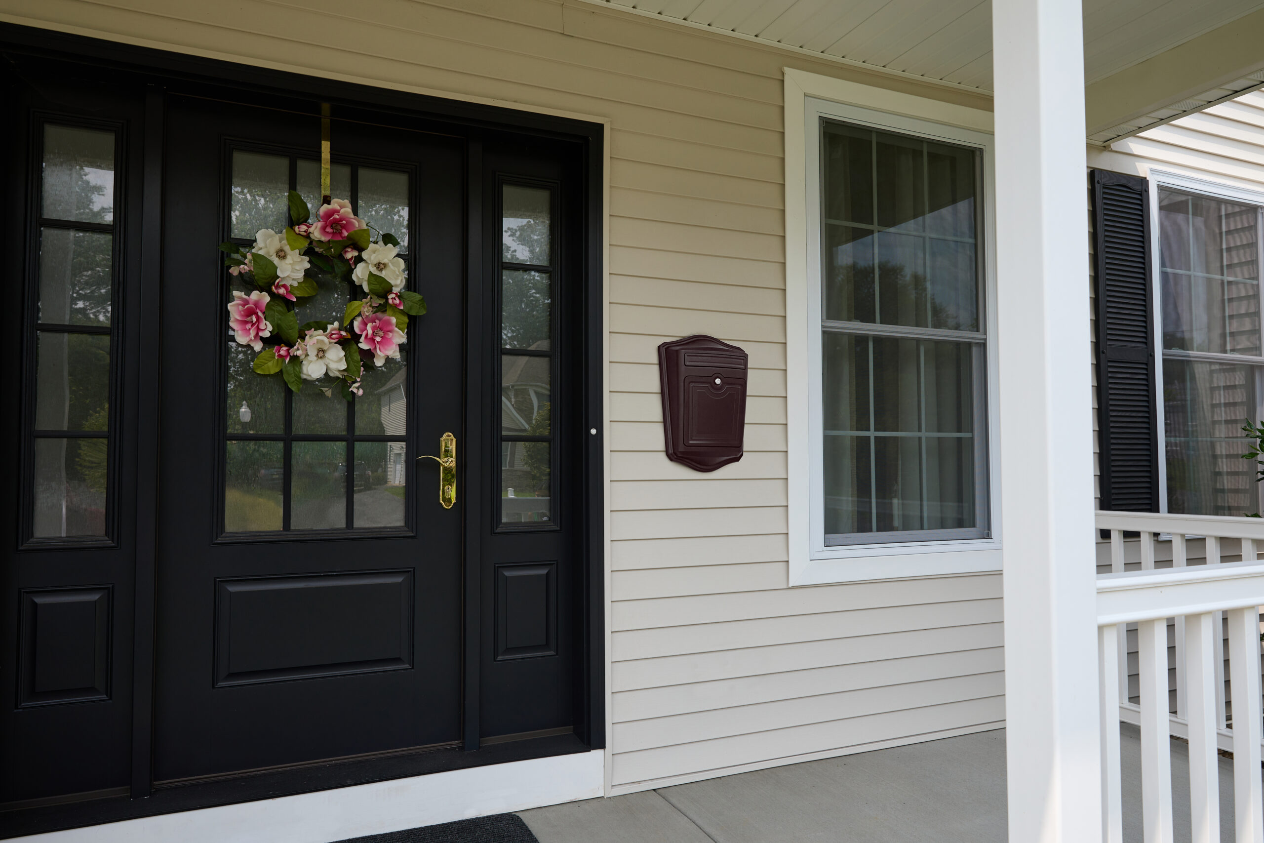 Black front door with glass panels and a pink‑white flower wreath; beige siding and a maroon mailbox on the wall nearby.