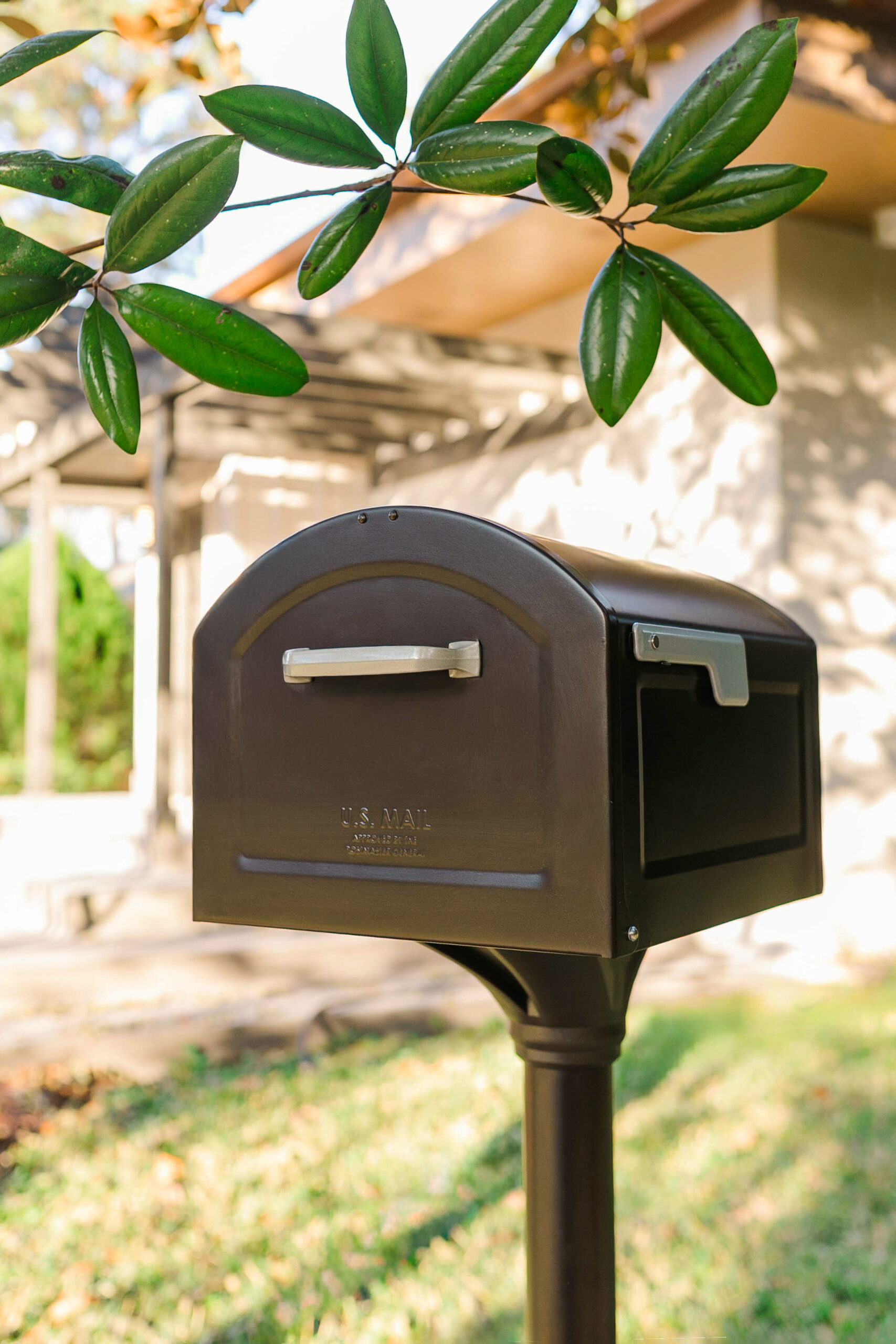 Brown U.S. mailbox on a post with green leaves overhead in a suburban yard