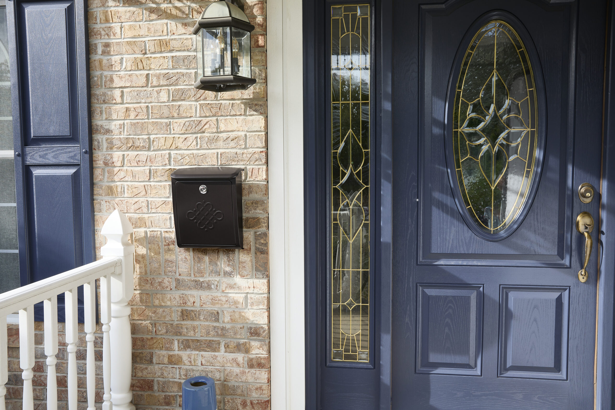 Blue front door with gold-edged oval glass, flanked by decorative sidelights, and a brick entryway with a wall lantern and black mailbox on the wall