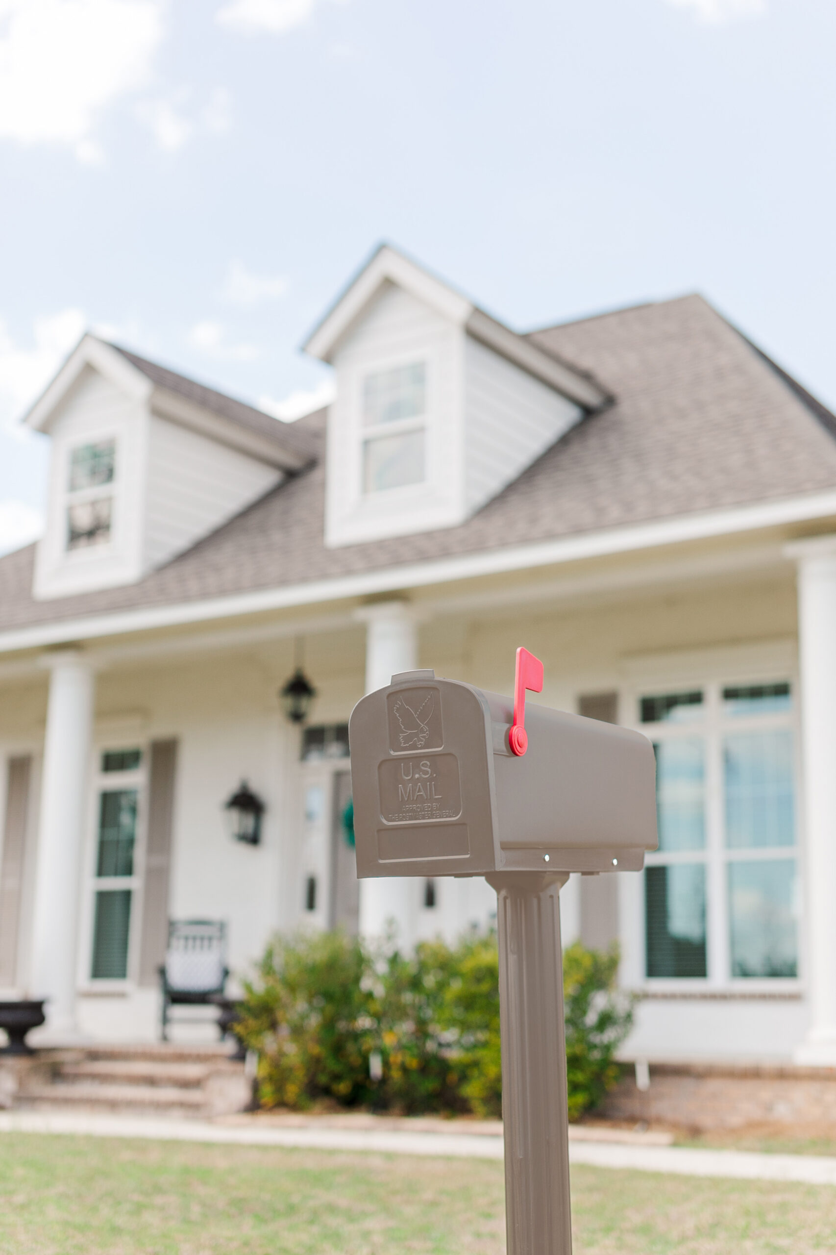 Gray USPS mailbox on a post with the pink flag raised in front of a two-story suburban home with dormer windows.