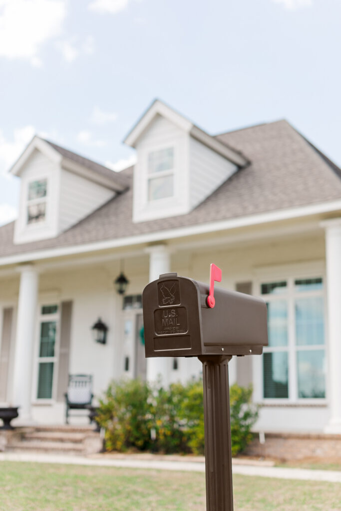 Brown US Mail mailbox on a post in front of a white house with dormer windows, suburban yard visible in background.