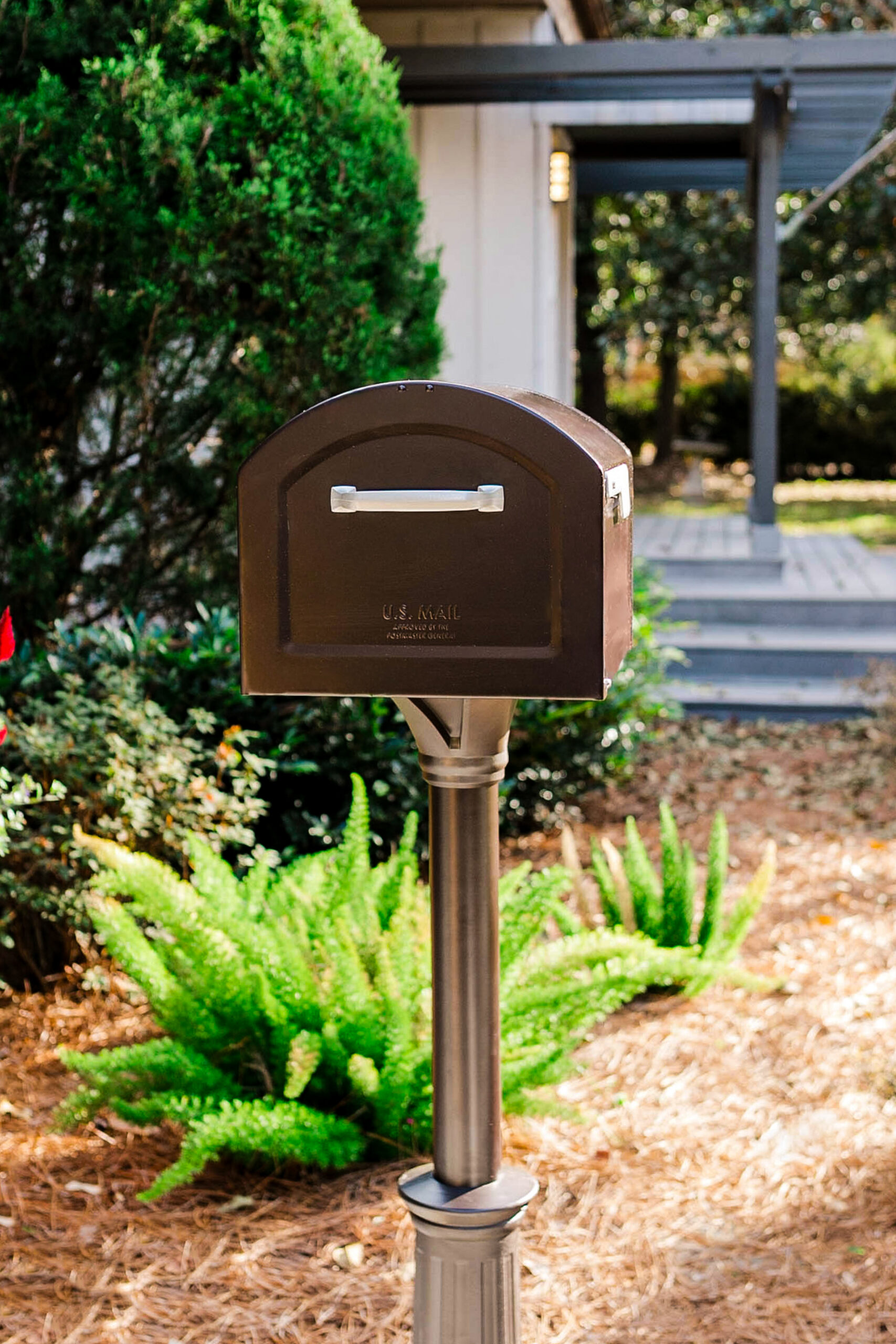 Brown US mail mailbox on a metal post in a landscaped yard with green shrubs and a porch in the background.