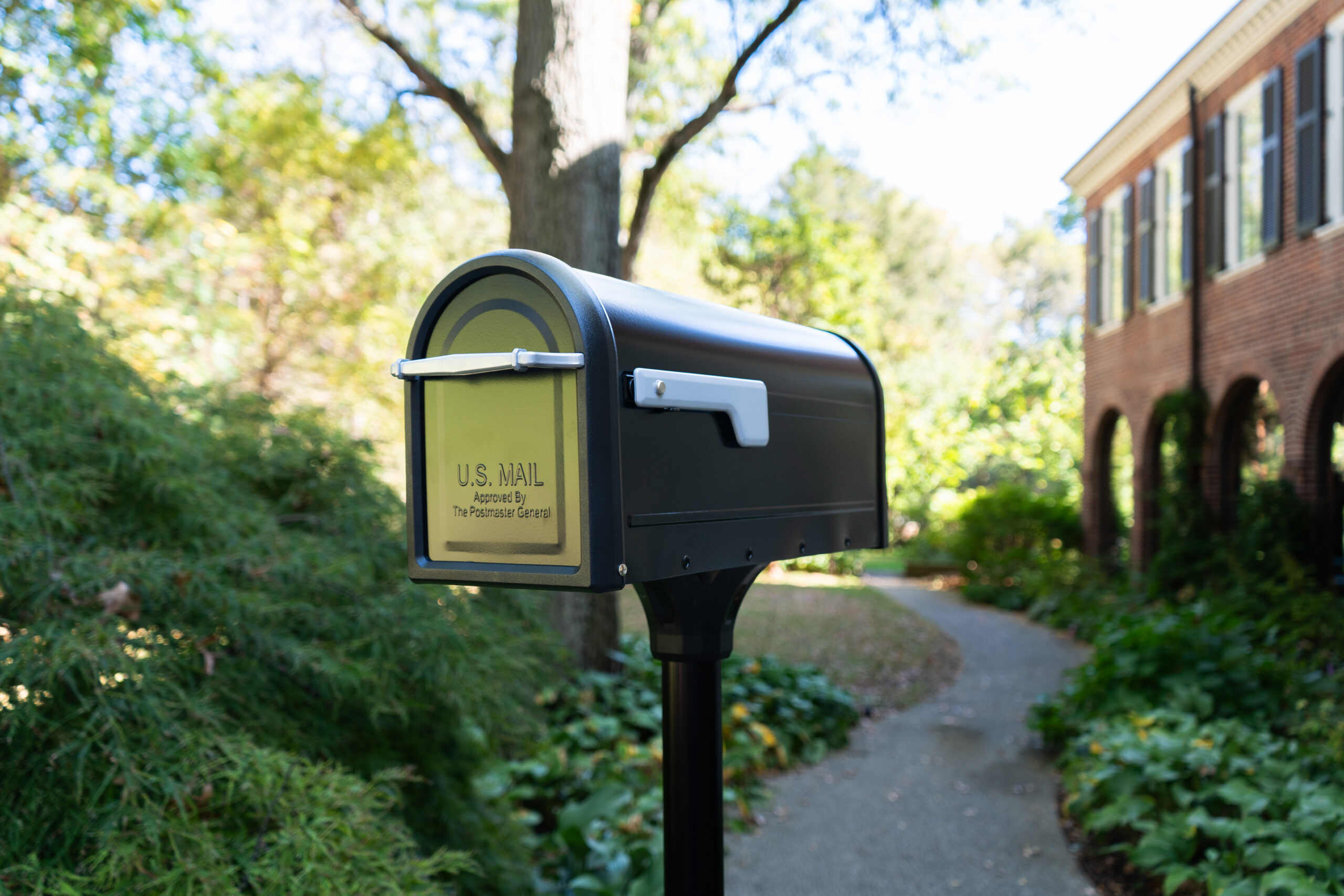 A dark mailbox on a black post beside a sidewalk with a brick building and green shrubs in the background.
