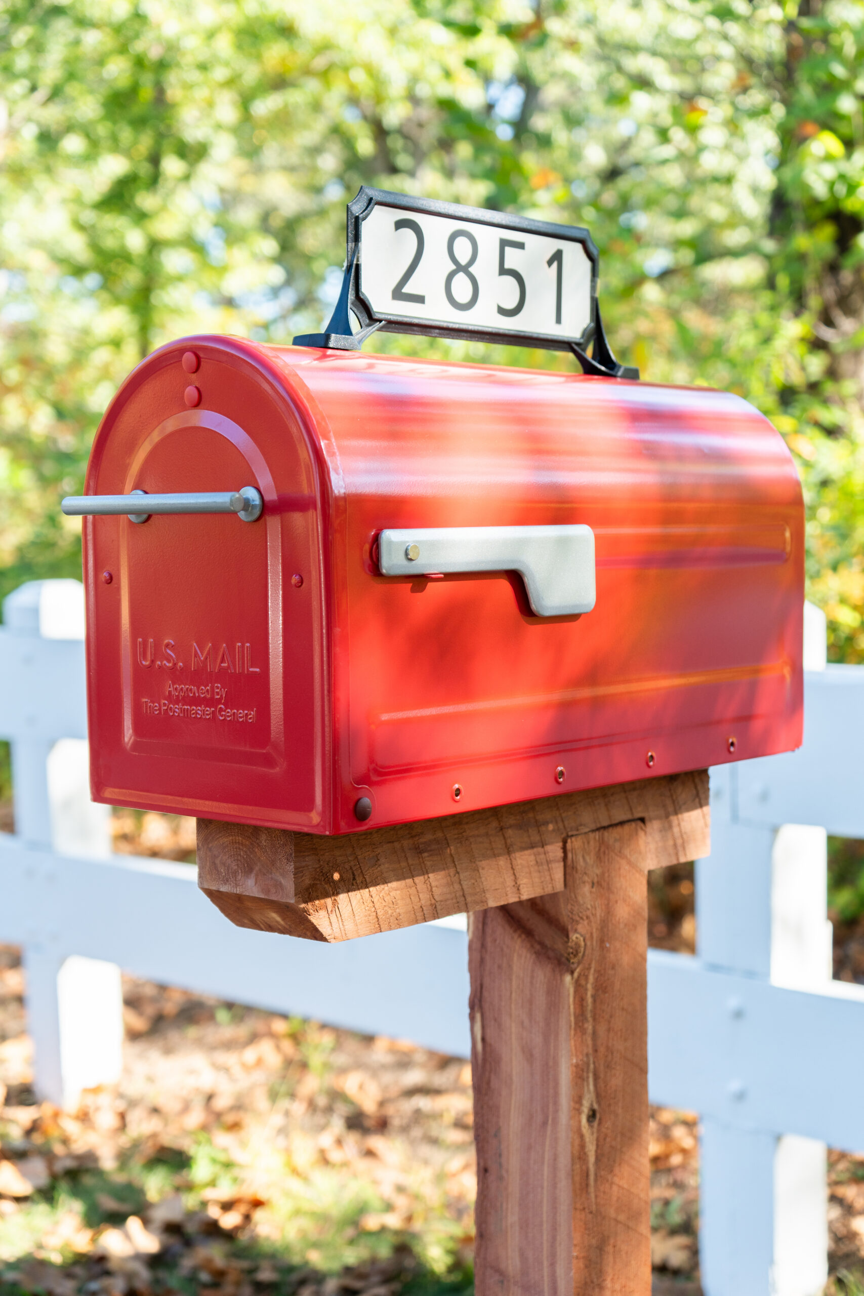 Bright red US mailbox mounted on a wooden post, displaying the number 2851 on a sign, with a white fence and greenery in the background.