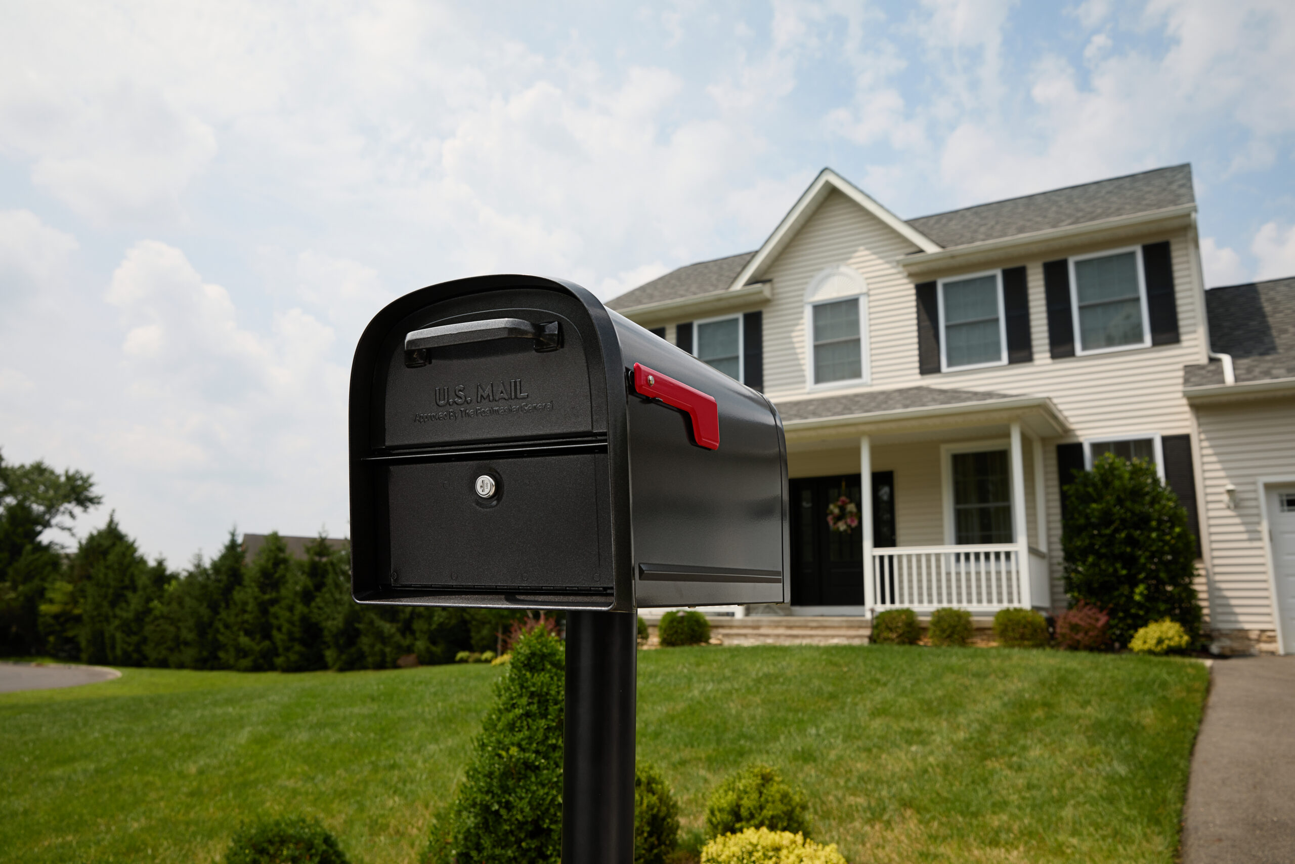 Black U.S. Mail mailbox with a red flag in the foreground, front yard of a two-story beige house with a porch and trimmed shrubs.