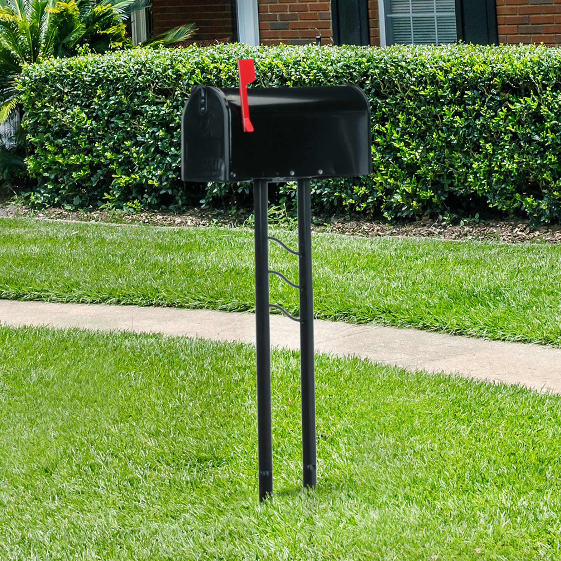 Black mailbox on a tall metal post with a red flag raised, in a suburban yard with green grass and hedges behind.