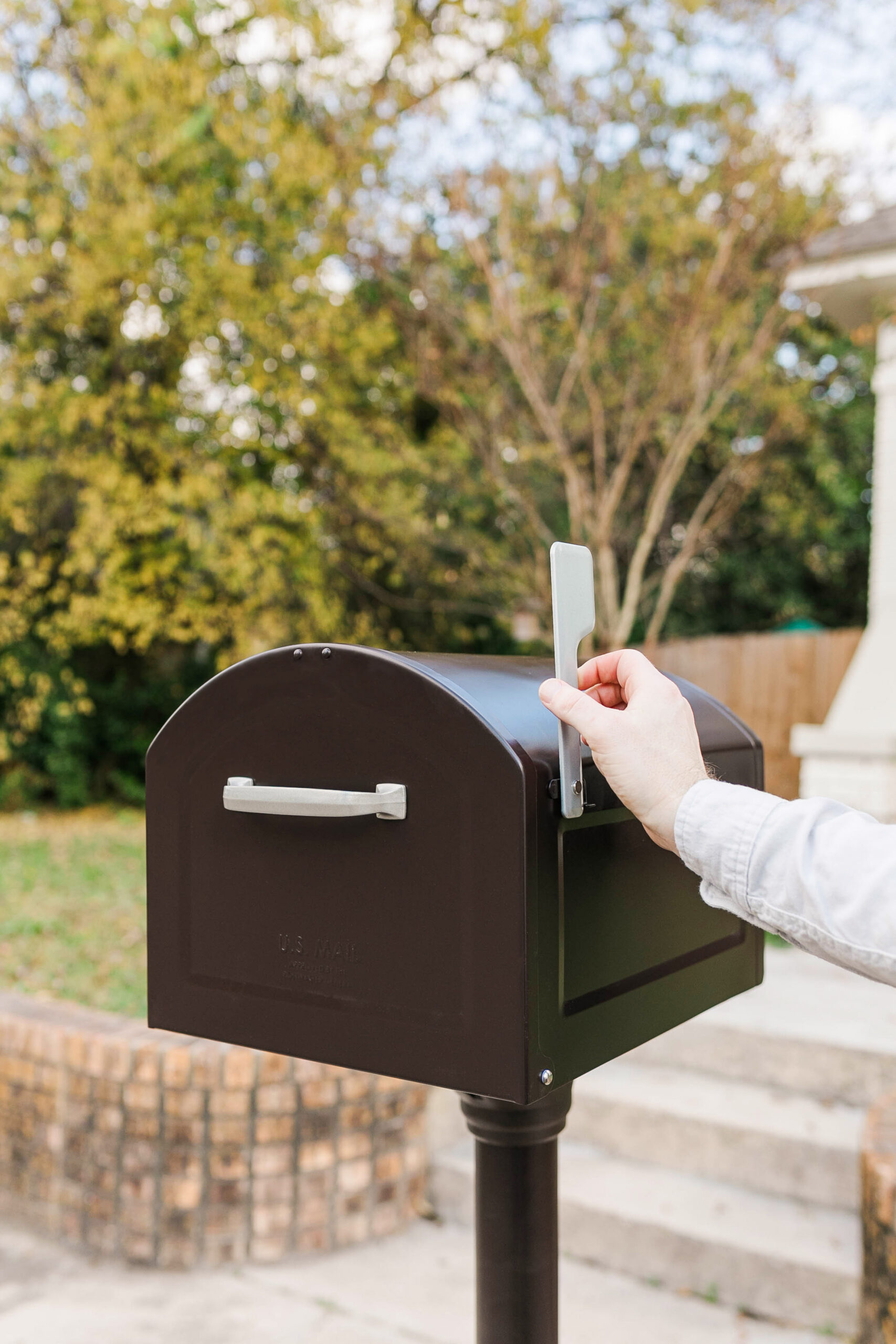Hands raise the silver mailbox flag on a dark brown curbside mailbox in a front yard.