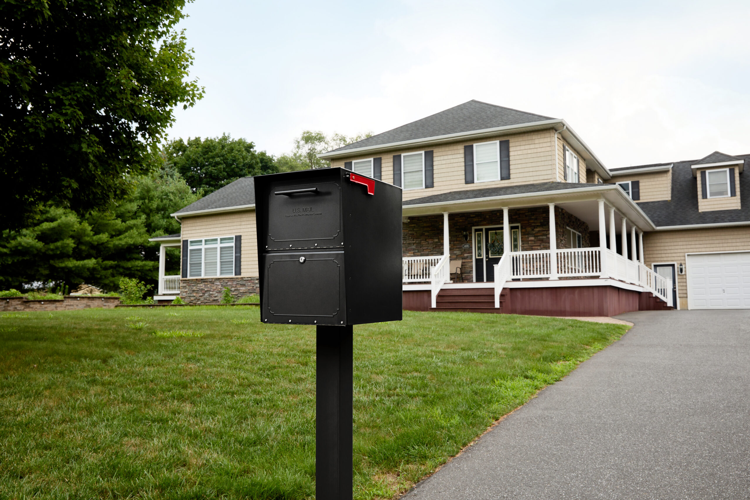 Black mailbox on a post in a green front yard with a two-story beige house and white porch in the background