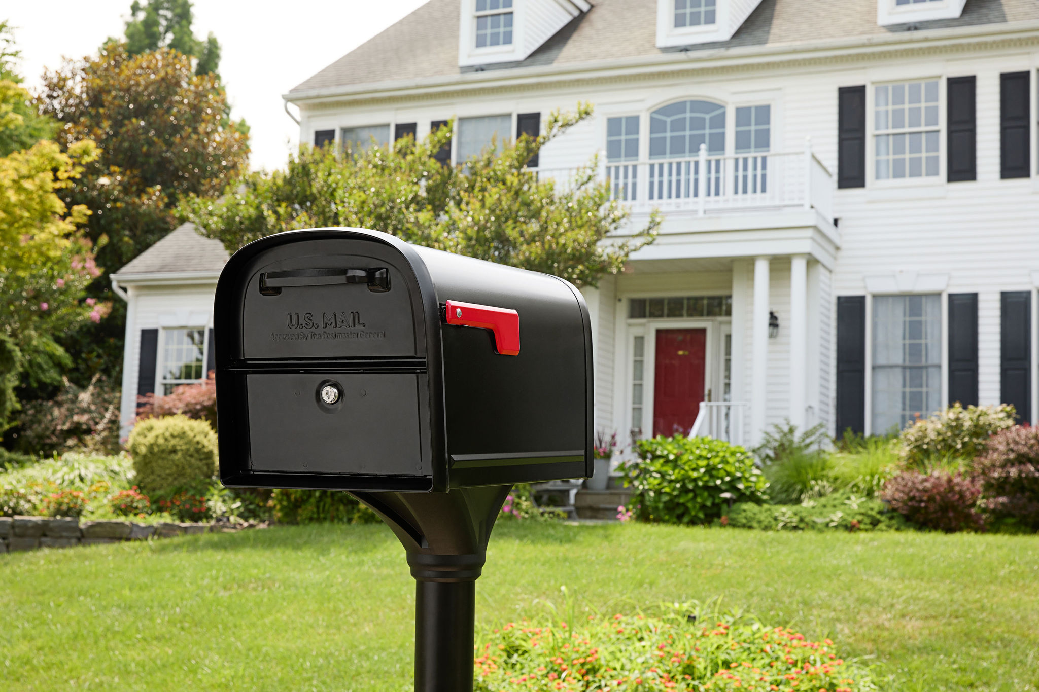 Black U.S. mailbox on a post in the front yard of a white two-story house with a red door and green lawn.