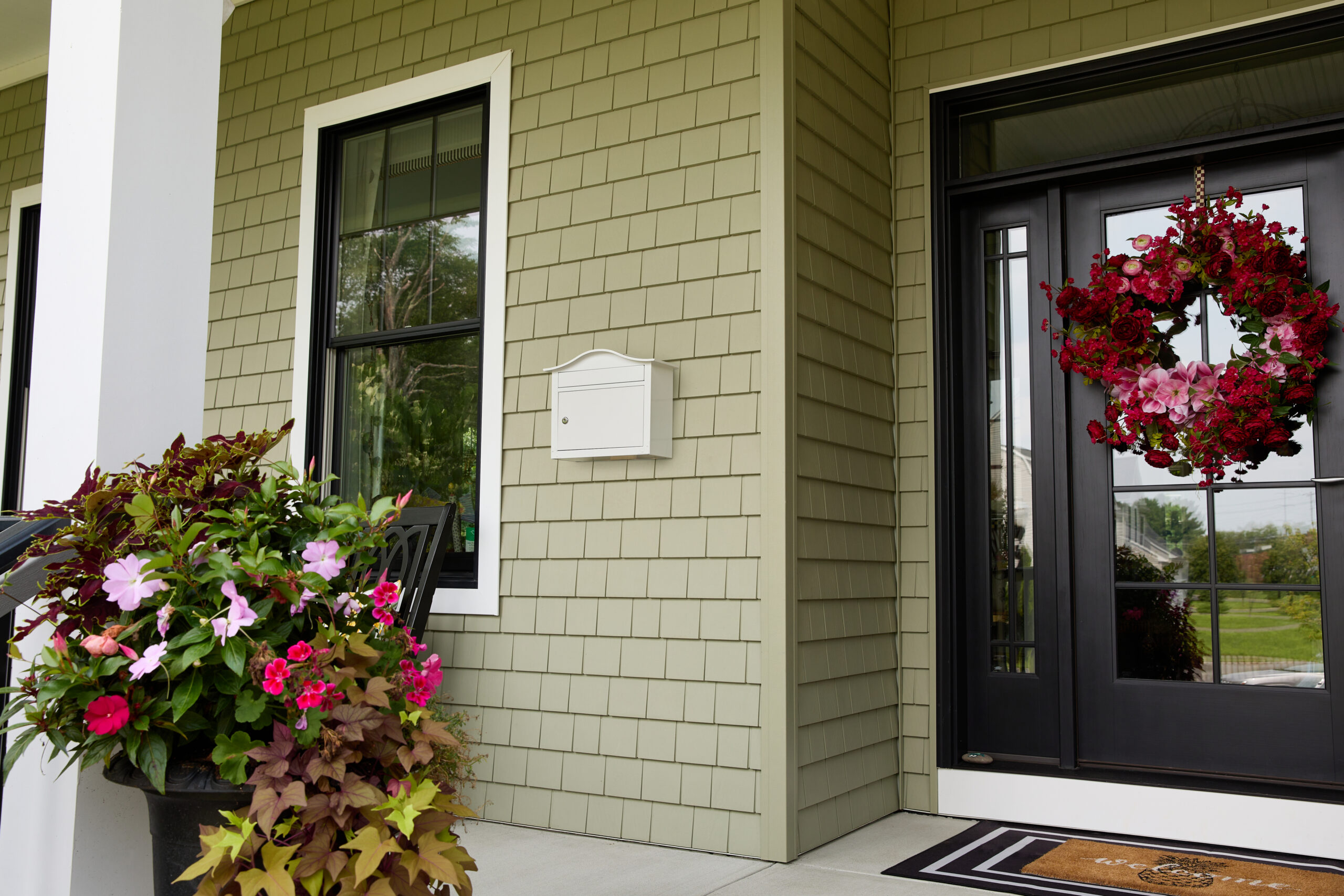 Front porch with a black front door and a vibrant red-pink flower wreath, beige siding, window, and a planter with colorful blooms next to the stairs