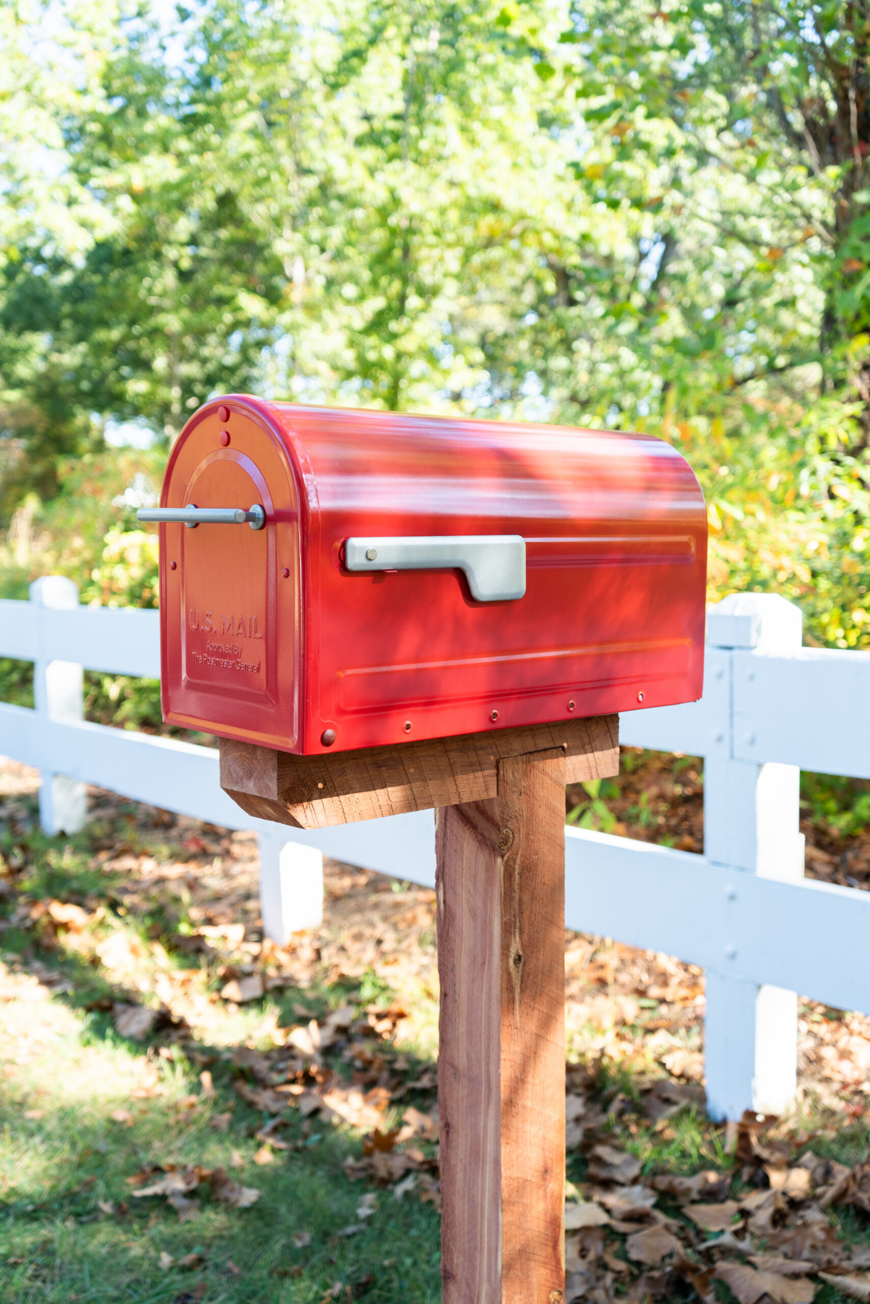 Red U.S. Mail mailbox on a wooden post beside a white fence in a sunlit yard with green trees behind it