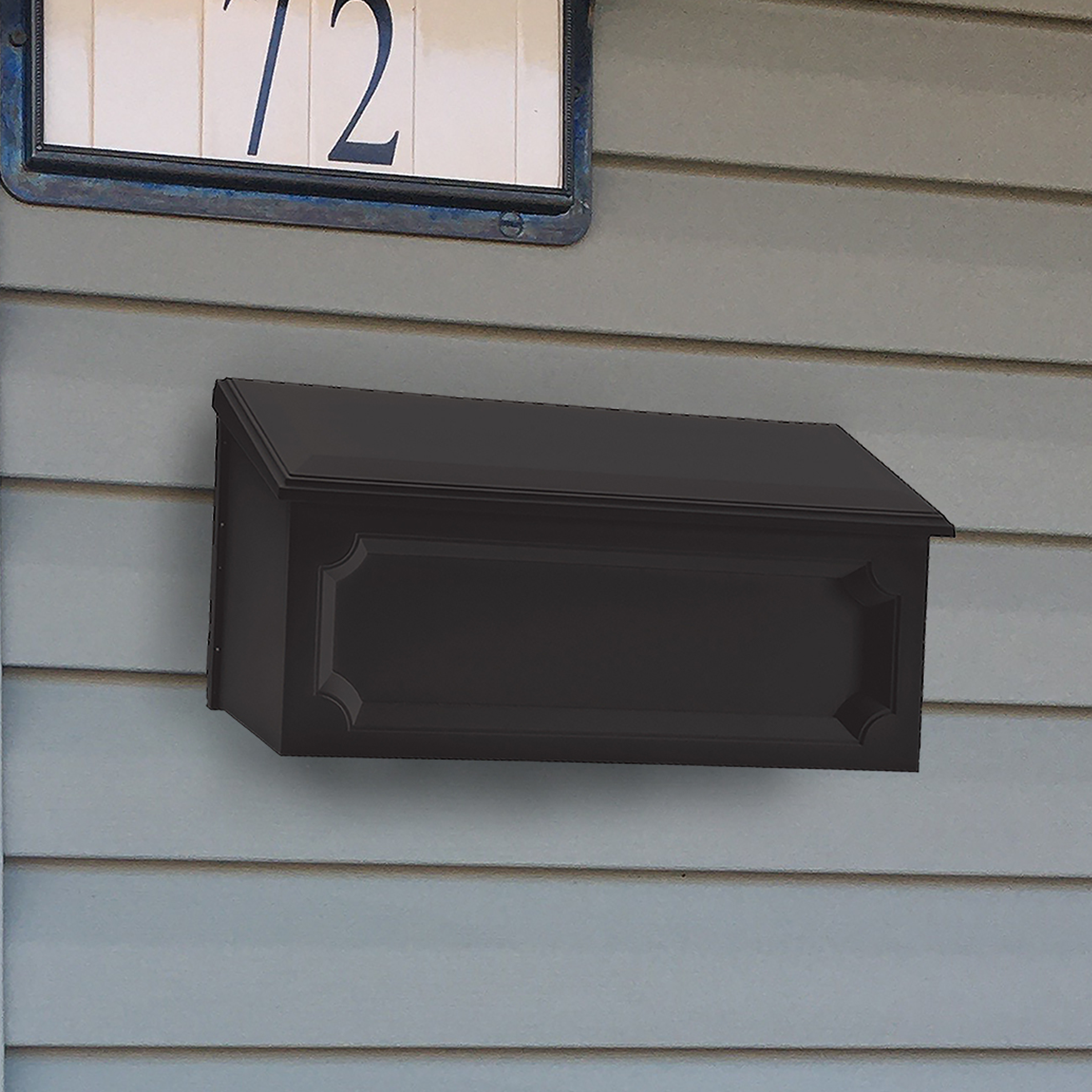 Black wall-mounted mailbox with decorative panels attached to gray siding beneath a window with the number 72 on a sign