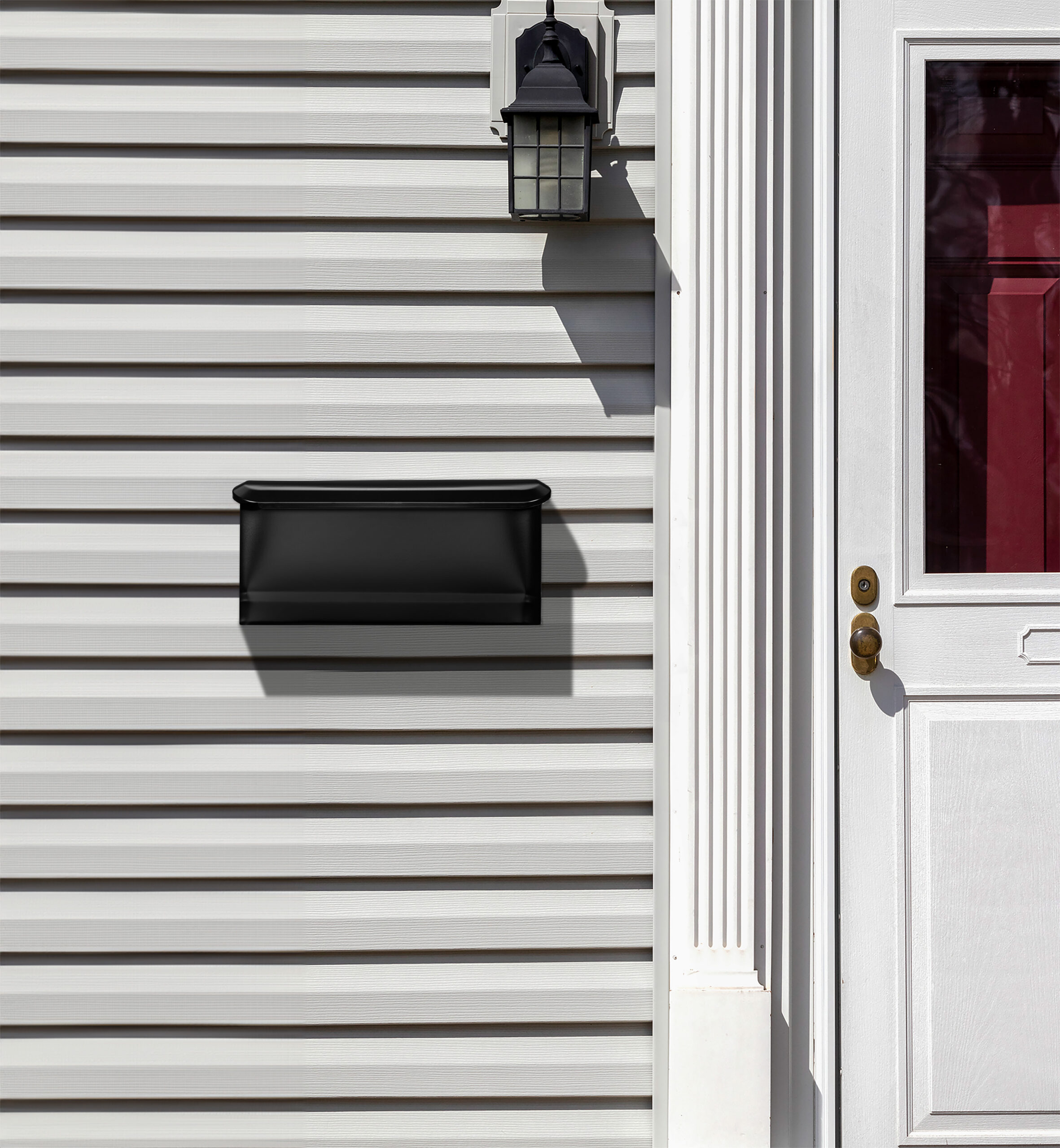 White front door on the right with brass knob, a black wall mailbox on the left, gray horizontal siding, and a black lantern light above.