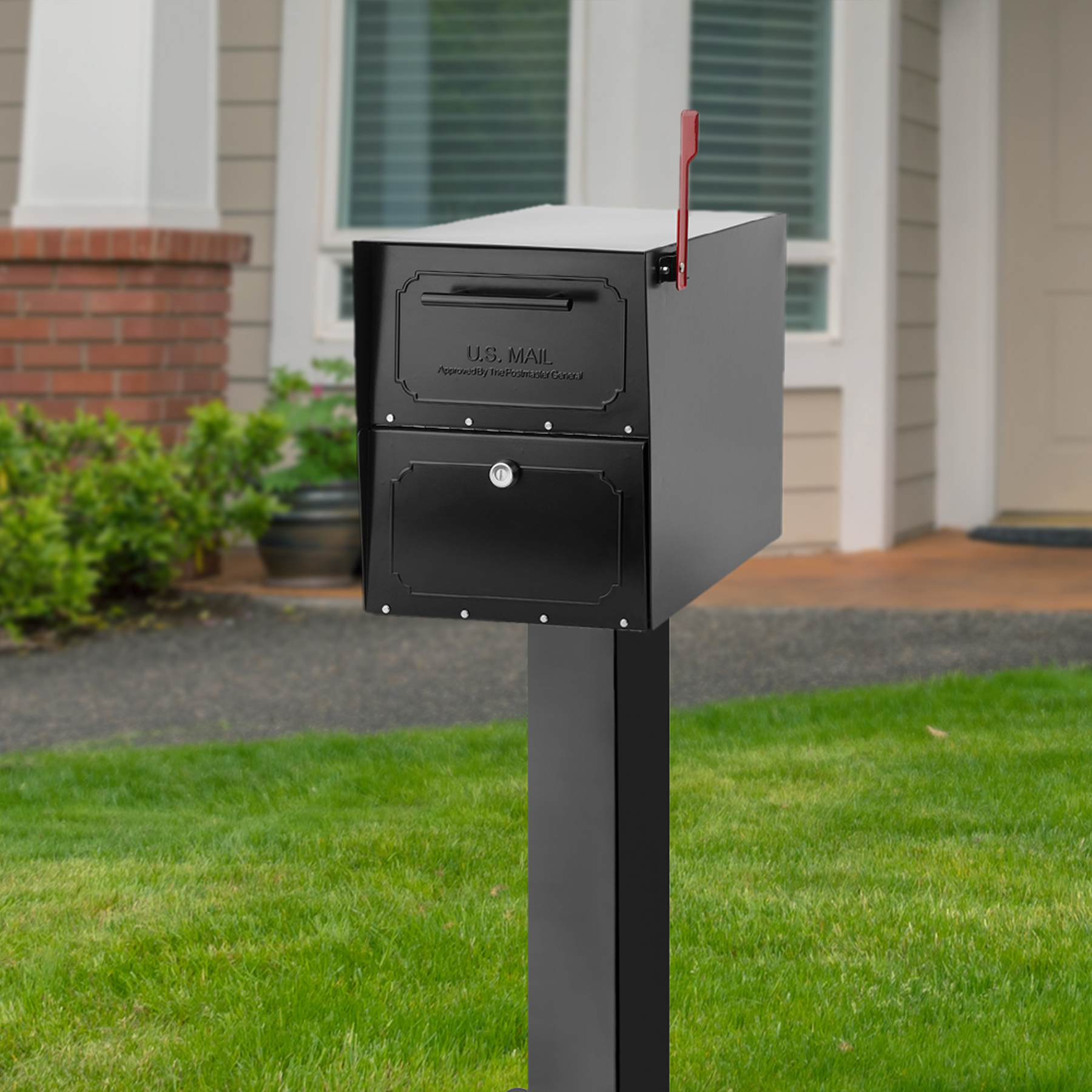 Black metal USPS mailbox with a raised red flag mounted on a tall post in a suburban front yard. Behind it are a brick and siding house, green lawn, and shrubs.