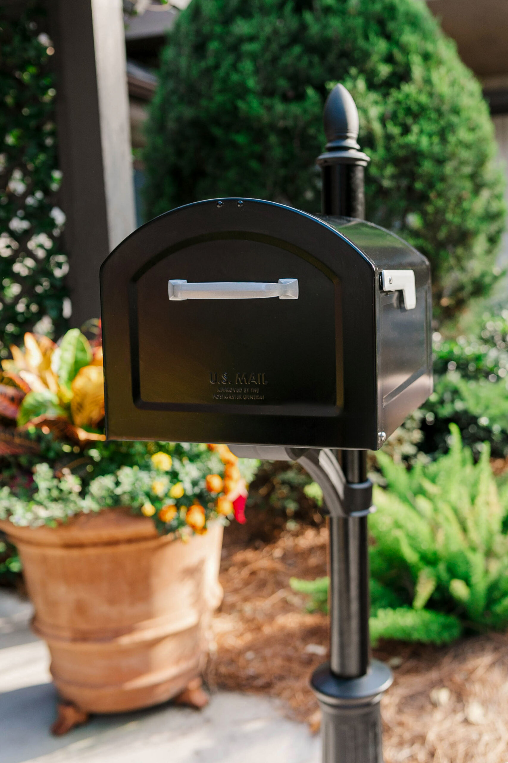 Black U.S. Mail mailbox mounted on a post in a garden with a potted plant nearby.
