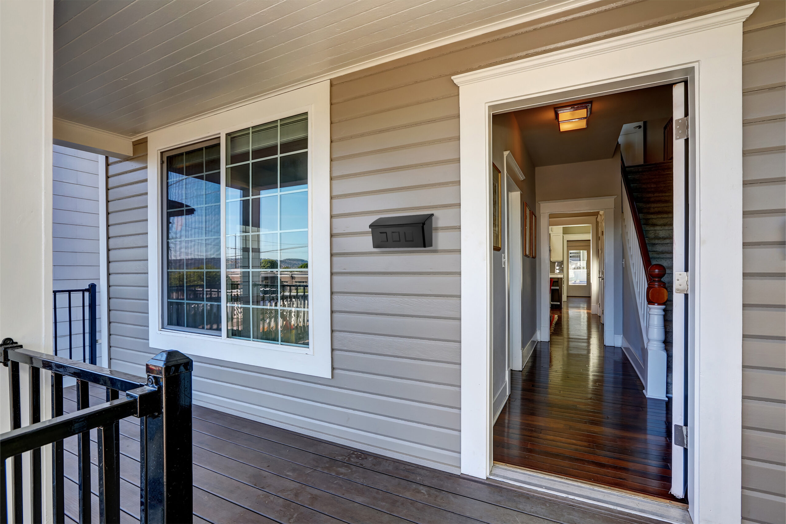 Open doorway to a home's hallway from a covered porch; a window with white trim and a black wall mailbox on beige siding,