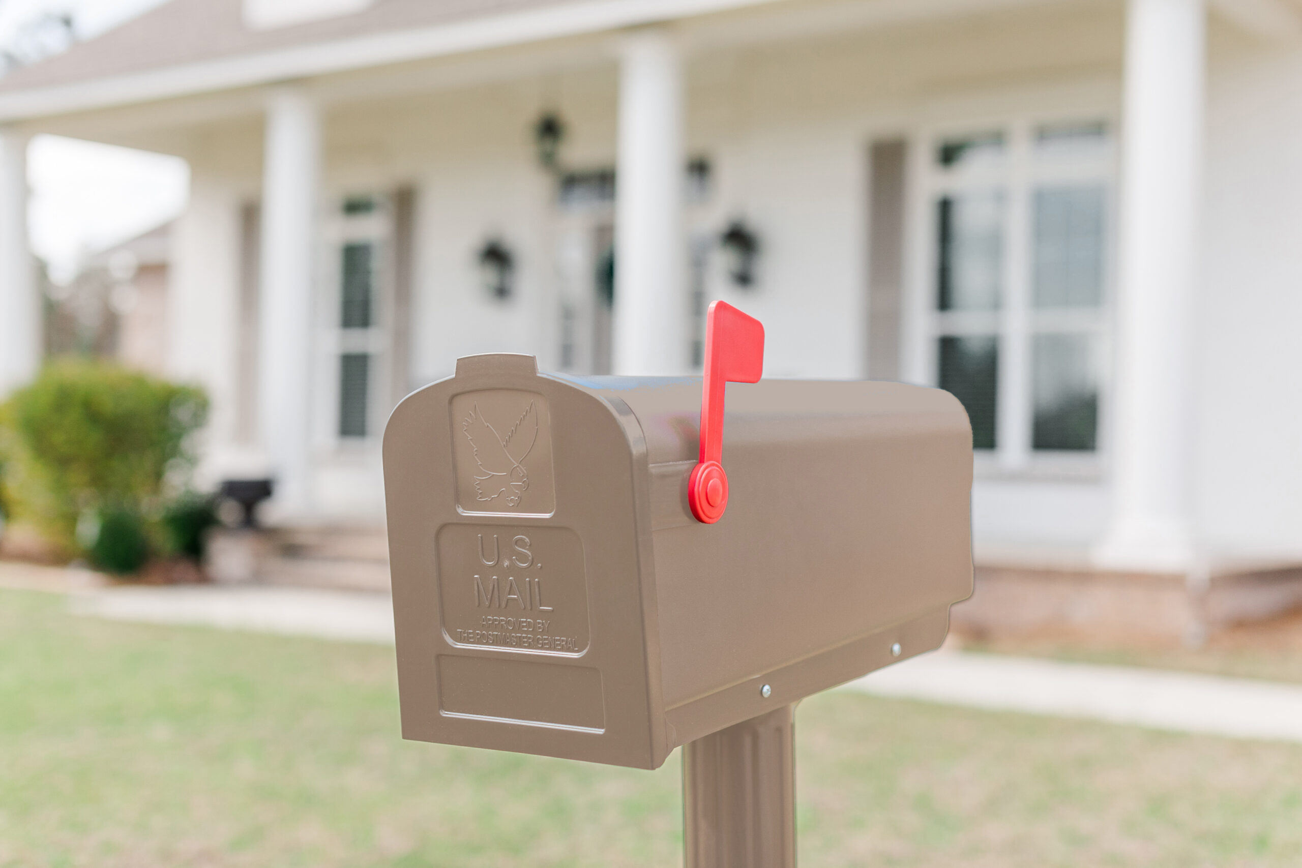 Brown curbside mailbox with a red flag raised, in front of a white house with columns (out of focus). It’s a residential mailbox on a post.