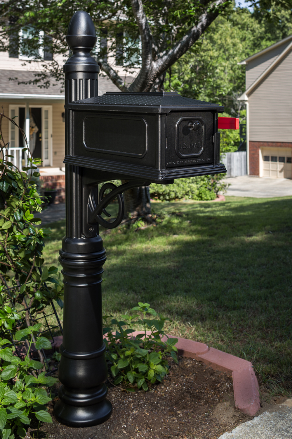 Black USPS mailbox mounted on a tall decorative post in a suburban front yard.