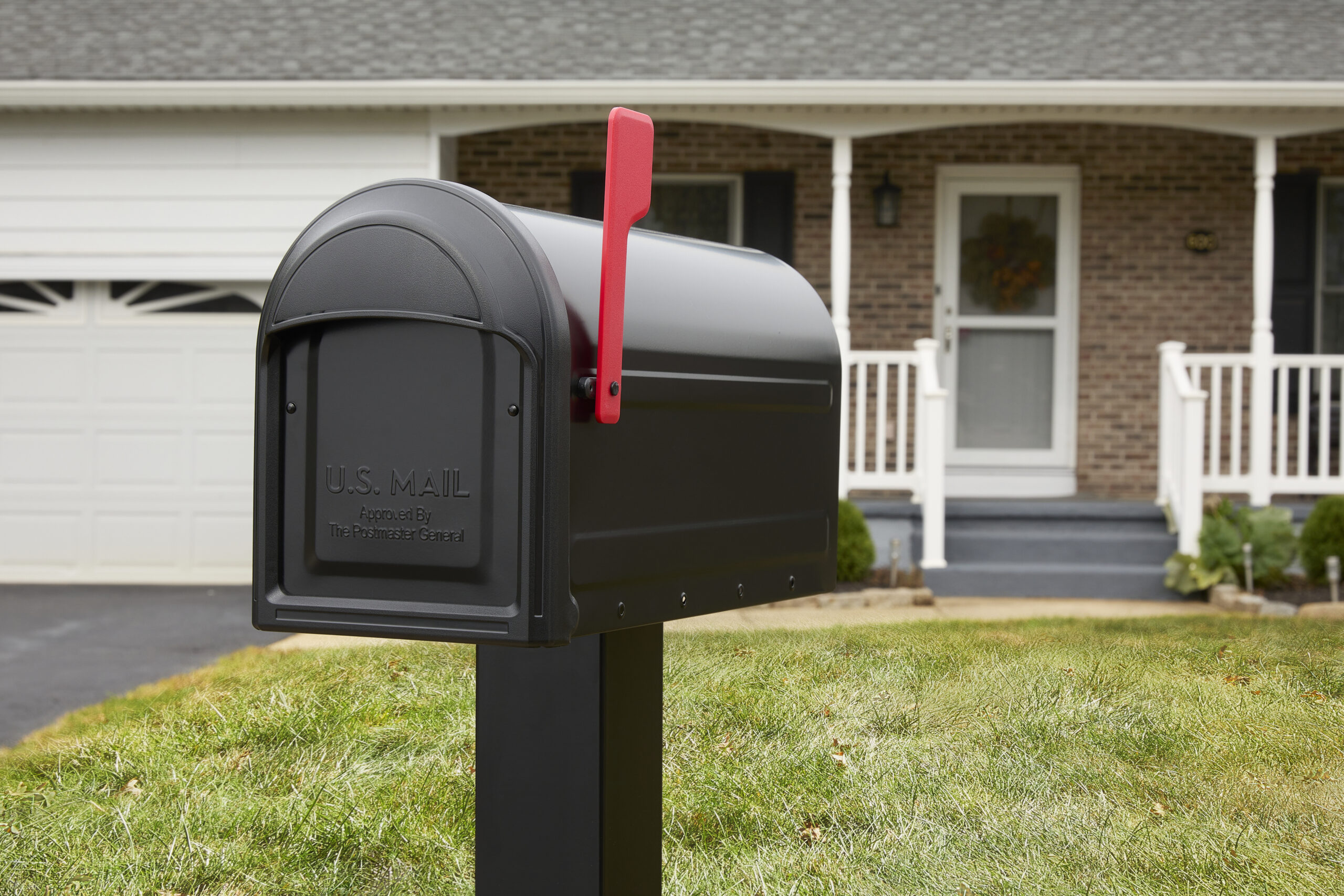 Black curbside mailbox on a post with the red flag raised, in front of a brick house with a white porch and garage in the background