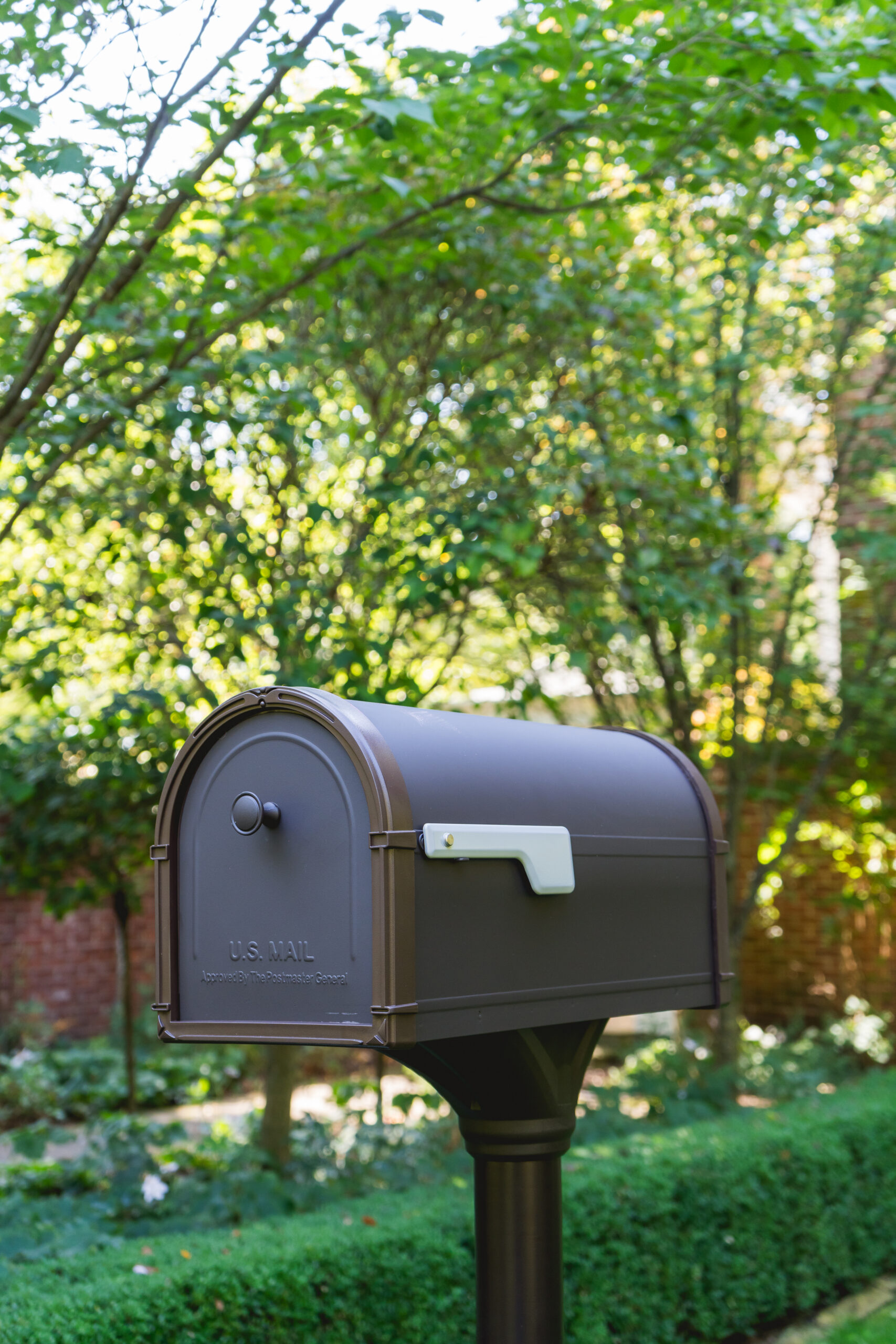 Grey curved-top U.S. Mail mailbox on a black post in a sunny garden setting. This is a residential curbside mailbox surrounded by green trees and shrubs.