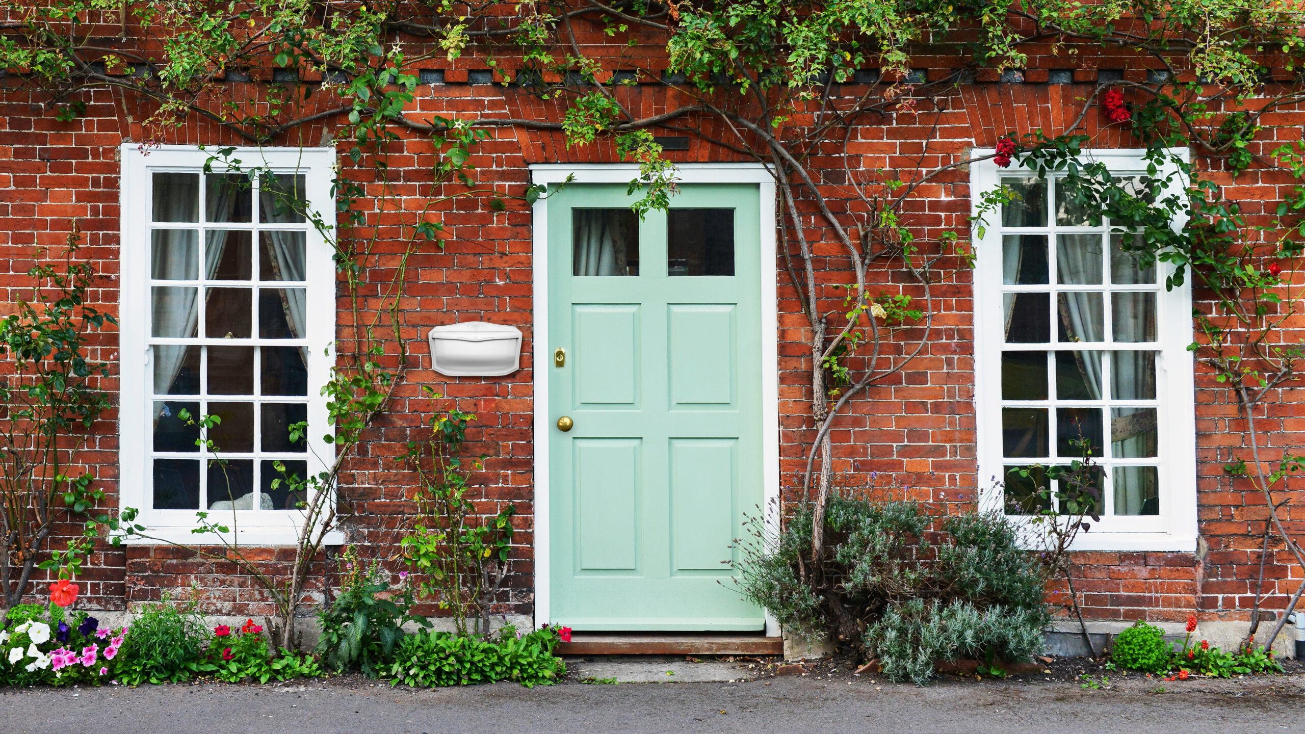 Brick townhouse facade with a mint green paneled door, two white-framed windows, and climbing vines; a small garden bed with colorful flowers sits along the foundation and a white wall mailbox is beside the door.