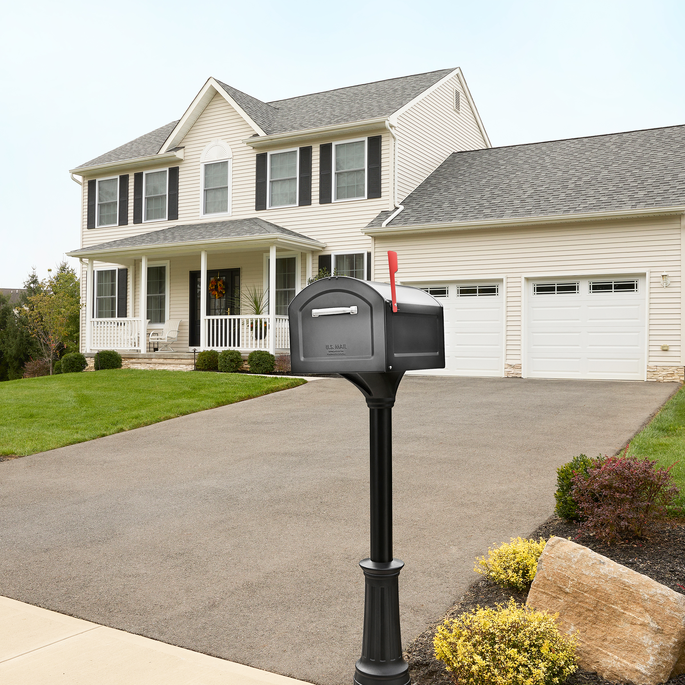 Black mailbox on a post in the driveway of a beige two-story house with a front porch and a two-car garage behind it.