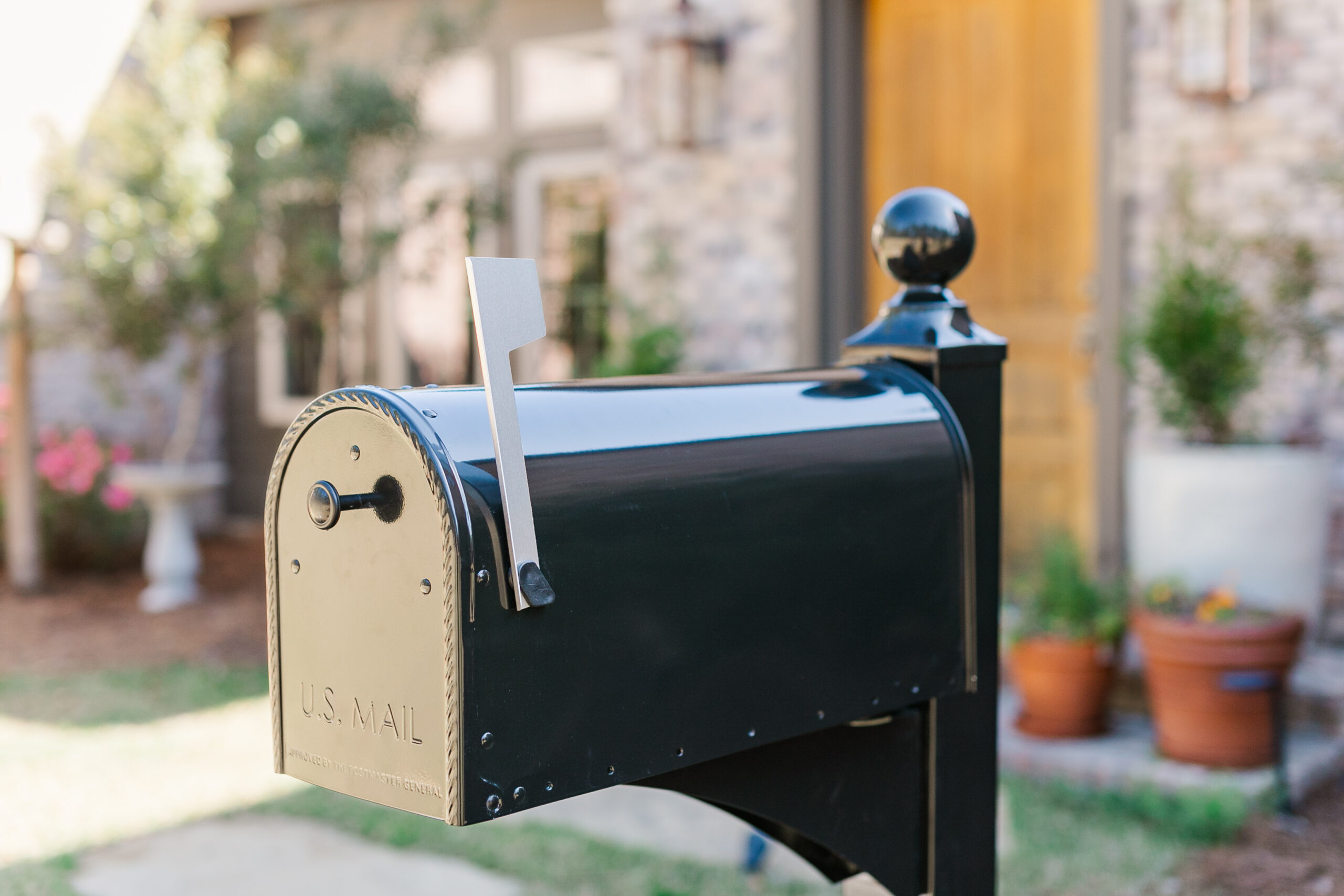 Black U.S. Mail mailbox with the flag raised, mounted on a post in a suburban yard with a blurred house and potted plants in the background.]}{