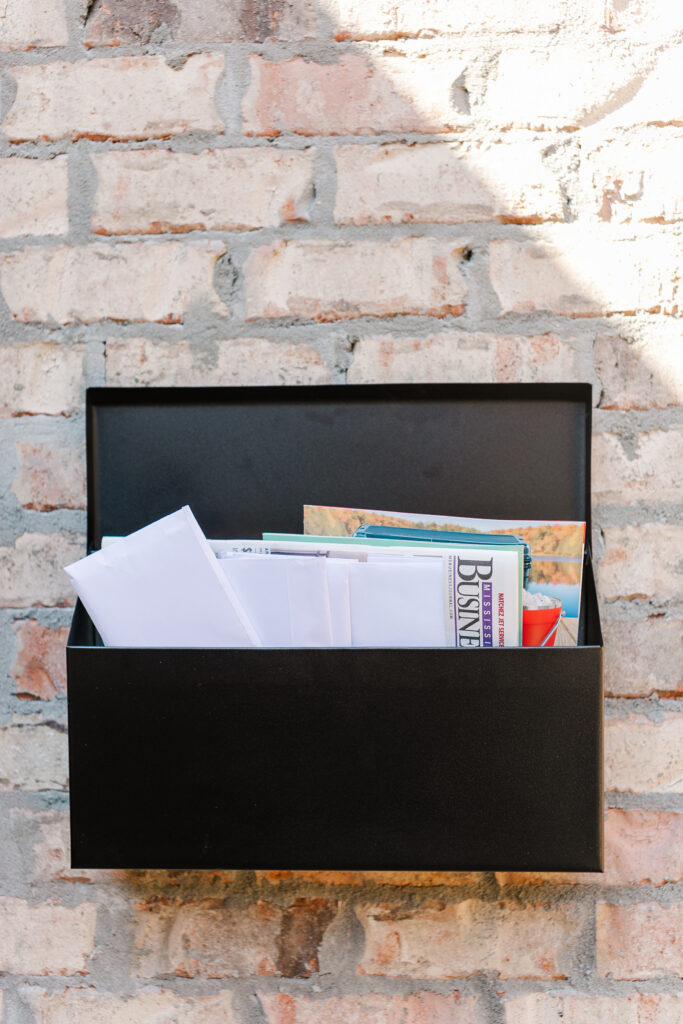 Open black wall-mounted mailbox filled with envelopes and a magazine against an exposed brick wall. extends a few inches from wall, sunlight casts shadow.