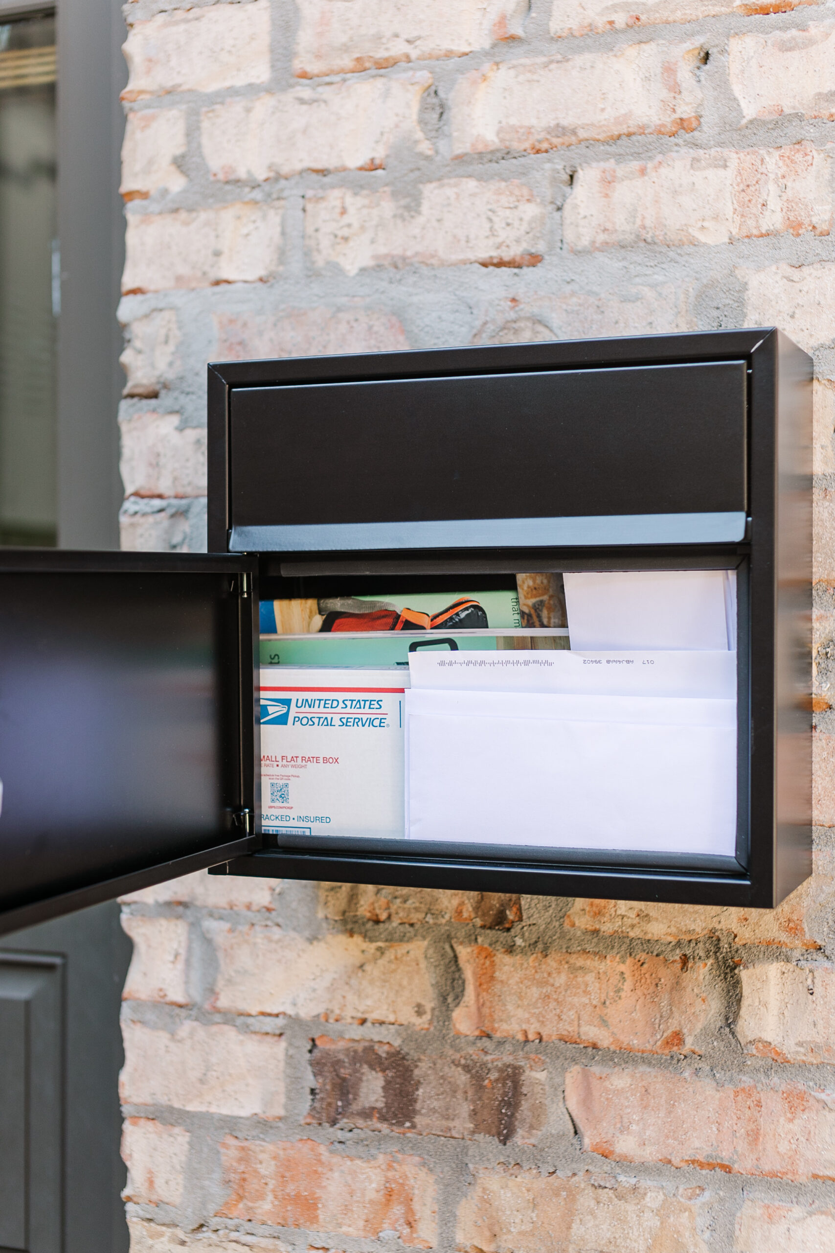 Black metal wall mailbox mounted on a brick exterior, with mail and papers visible inside and USPS materials showing in the lower compartment.