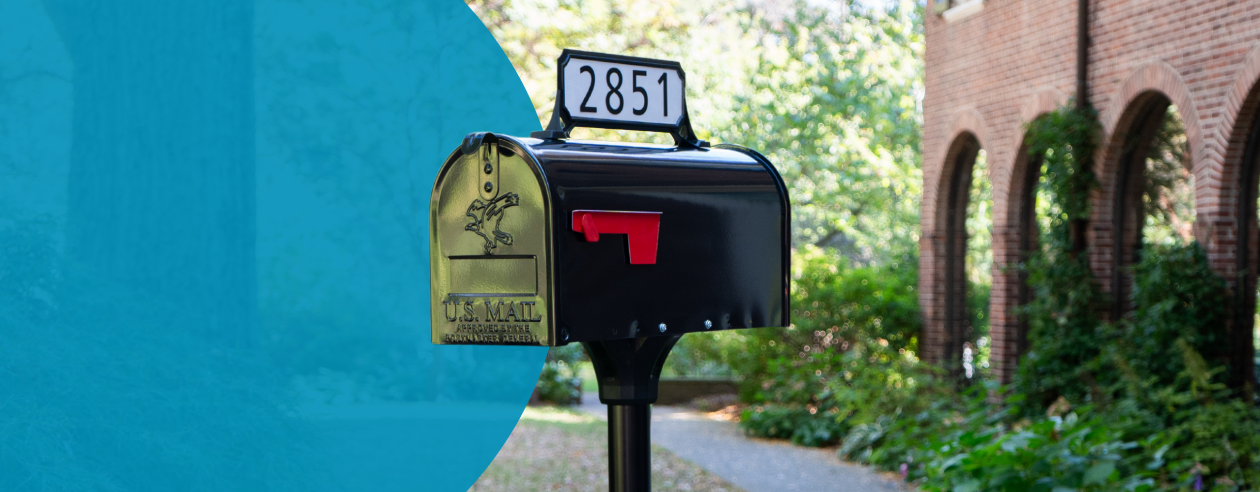 Black curbside mailbox with a raised red flag and the number 2851 on top, next to a brick building with arches in the background