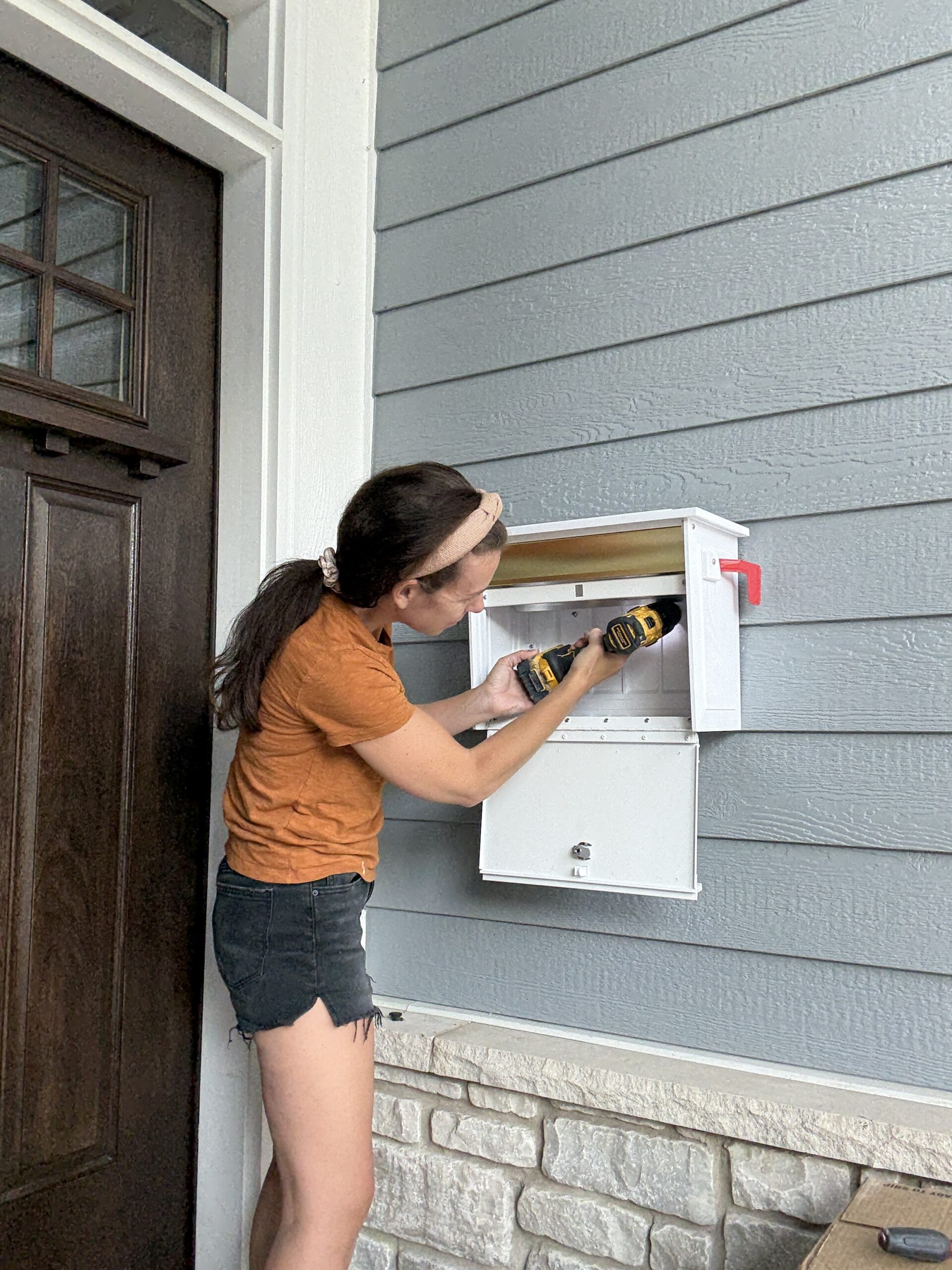 Woman installs a white wall-mounted mailbox/box beside a front door using a drill outside a house with blue siding.